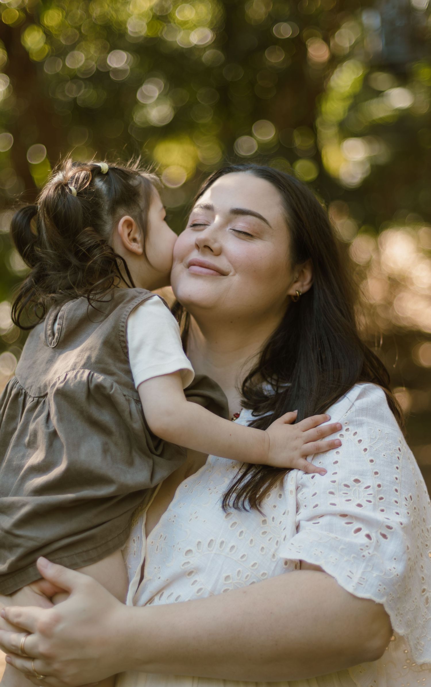 Mum smiling as her young daughter cuddles and kisses her during an outdoor family session.