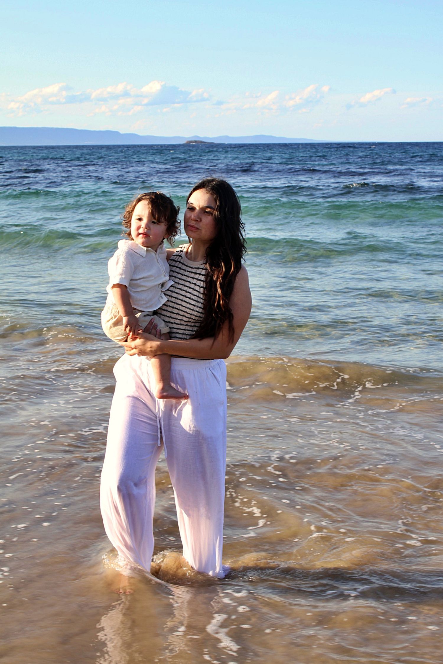 Mother holding her son, both looking at the camera smiling with the ocean behind them during a beach motherhood session.