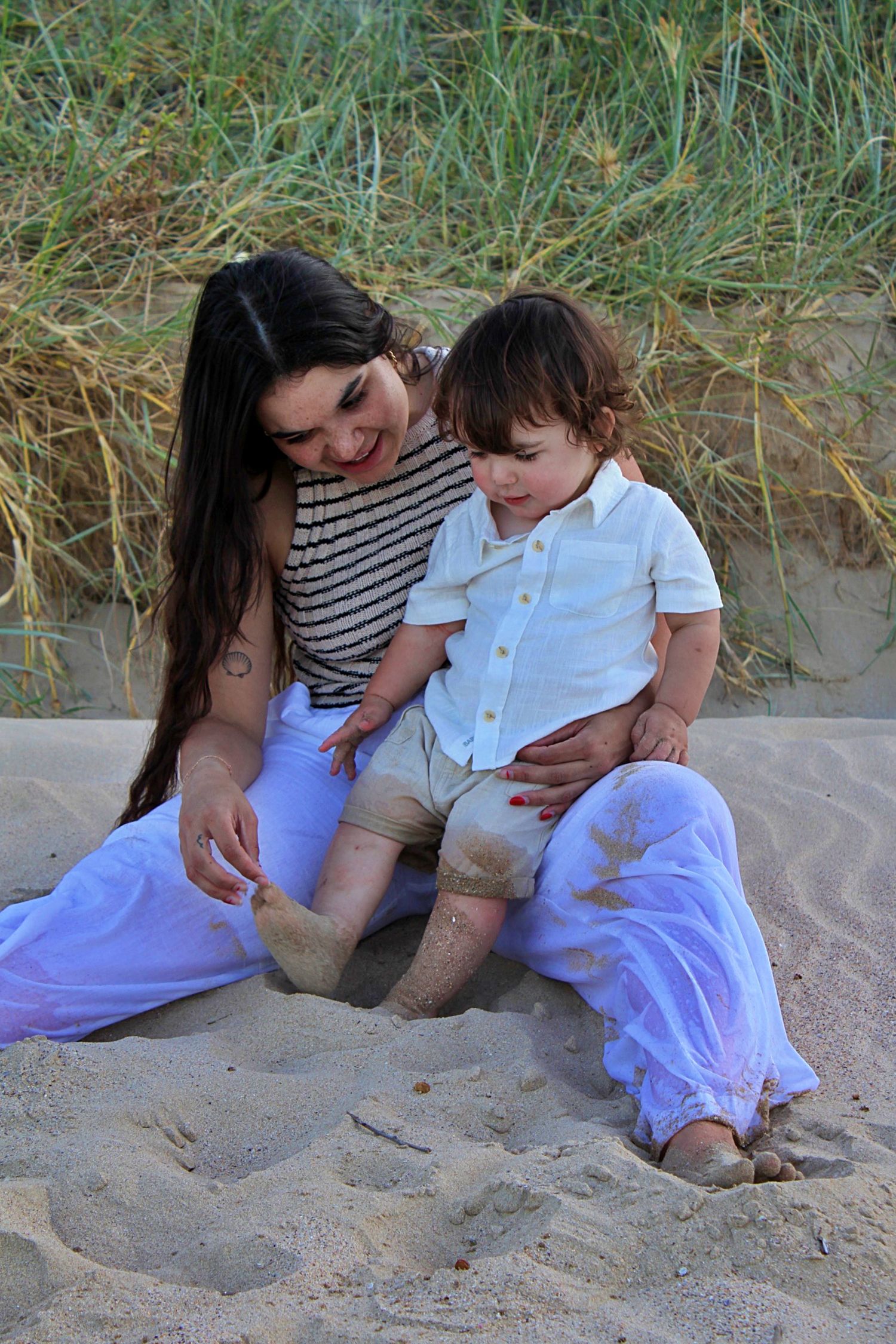 Motherhood session showing a mother and her son sitting on the beach with grassy lands in the background, playing with the sand together.