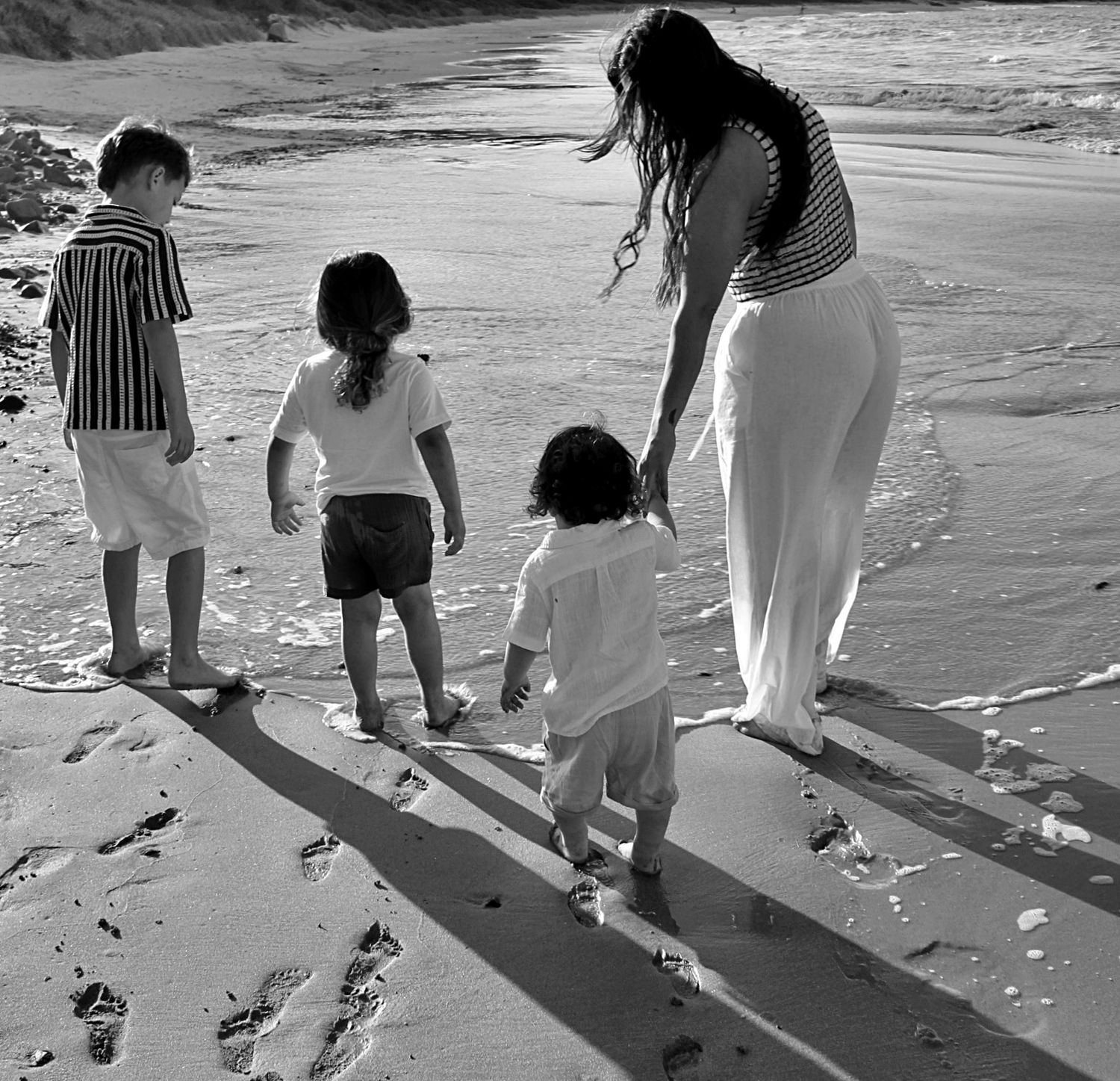 Mother walking hand in hand with her three boys along the beach, photographed from behind in natural light.