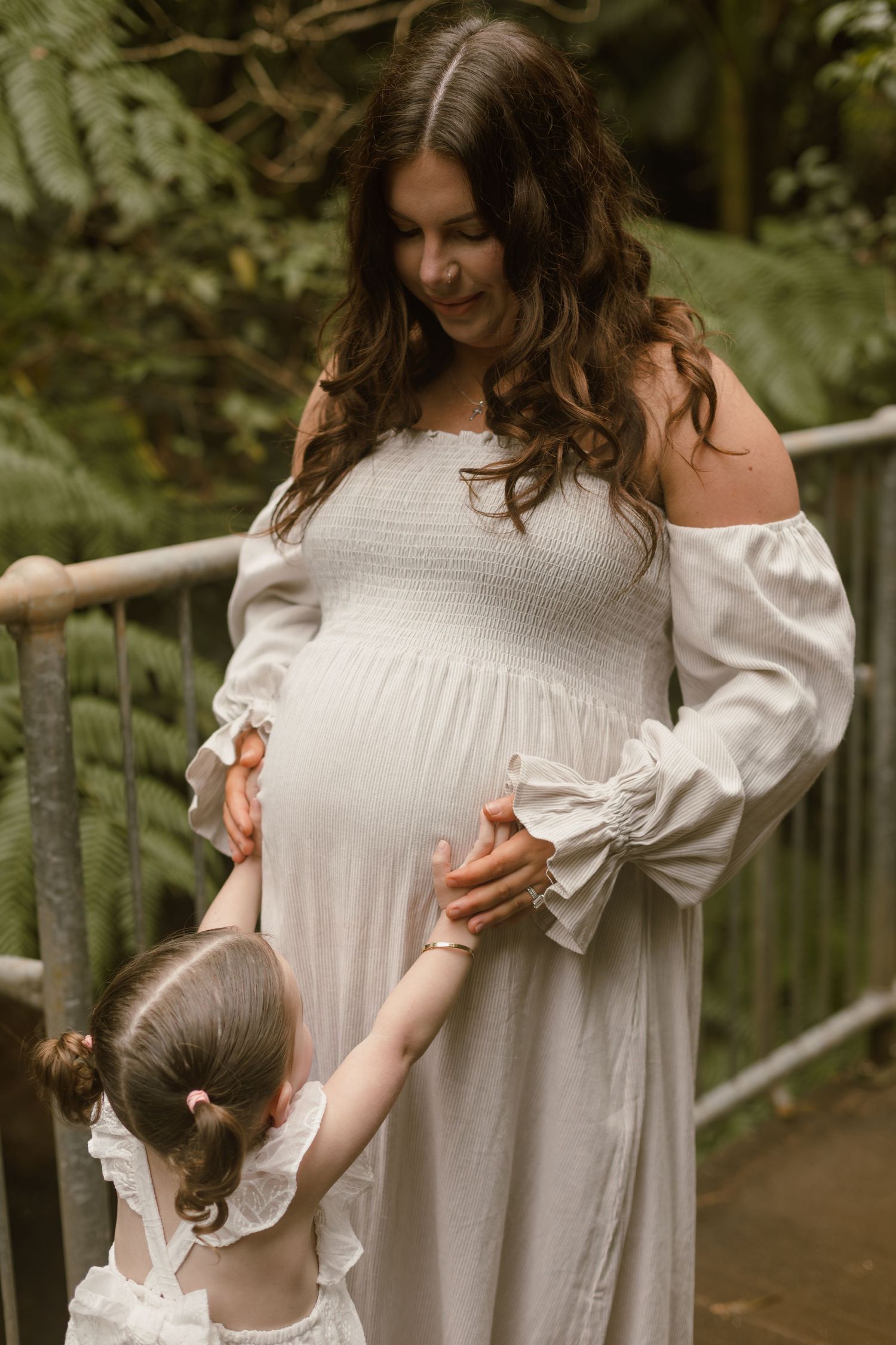 Young daughter reaching up to touch her mum’s baby bump during an outdoor family maternity photography session.