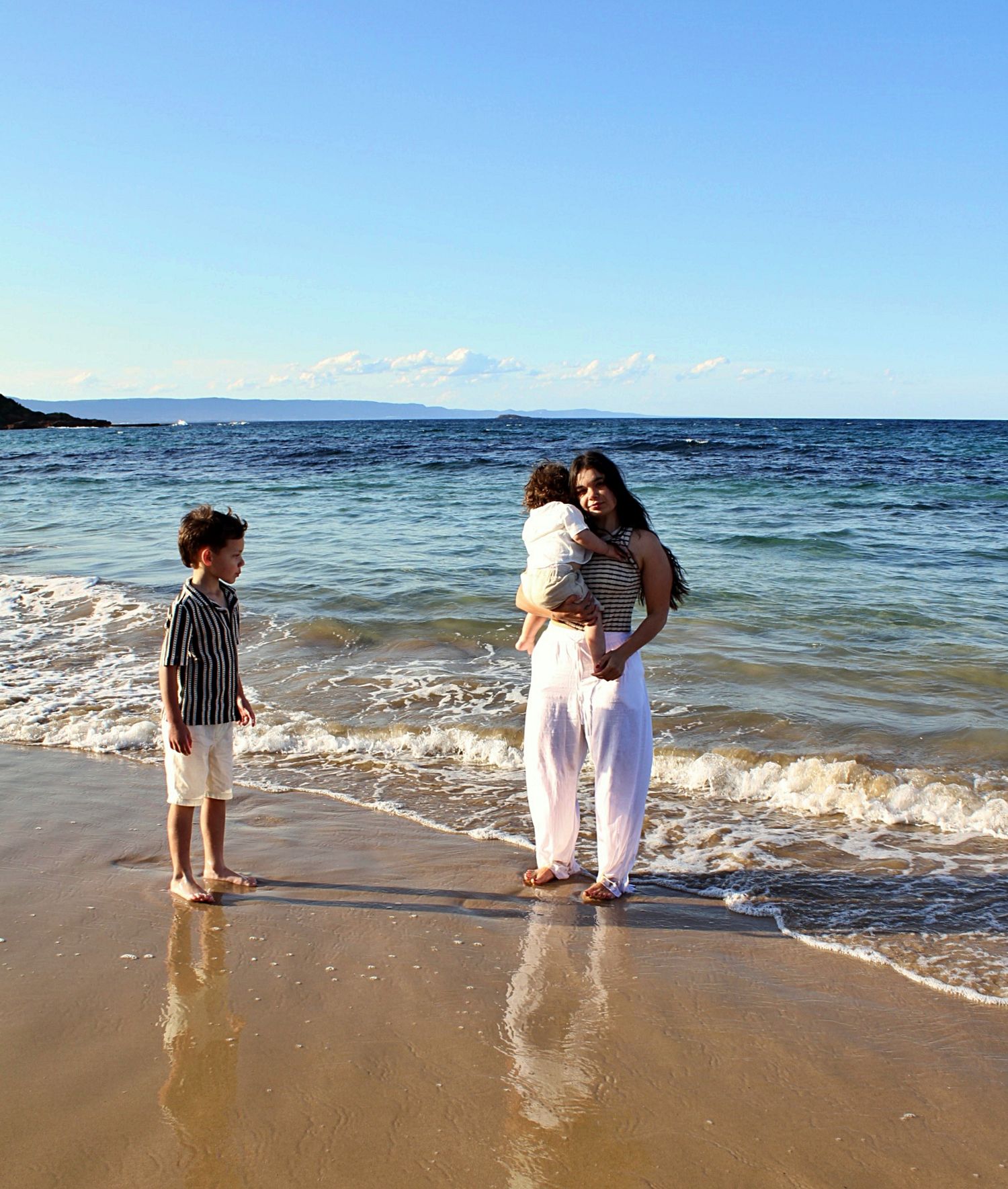 Candid moment of a mother holding her youngest son for a cuddle while another son watches by the shoreline during a beach motherhood session