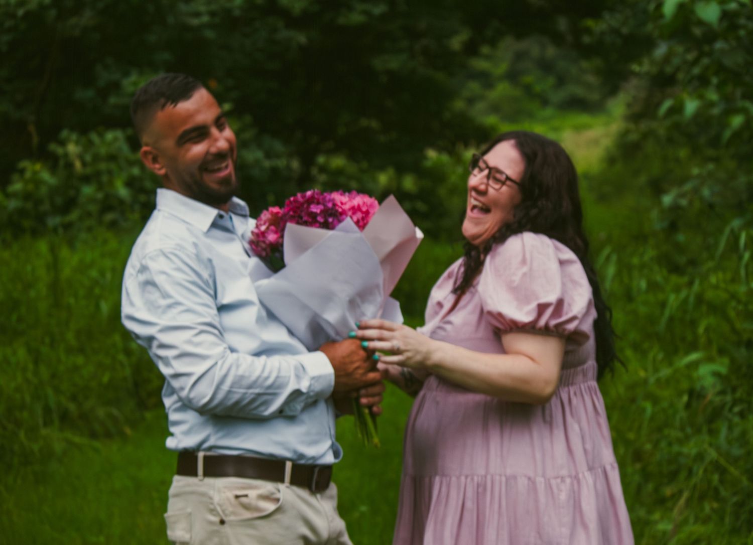 Couple laughing together during their gender reveal, holding a bouquet of pink flowers, celebrating the upcoming arrival of their baby girl
