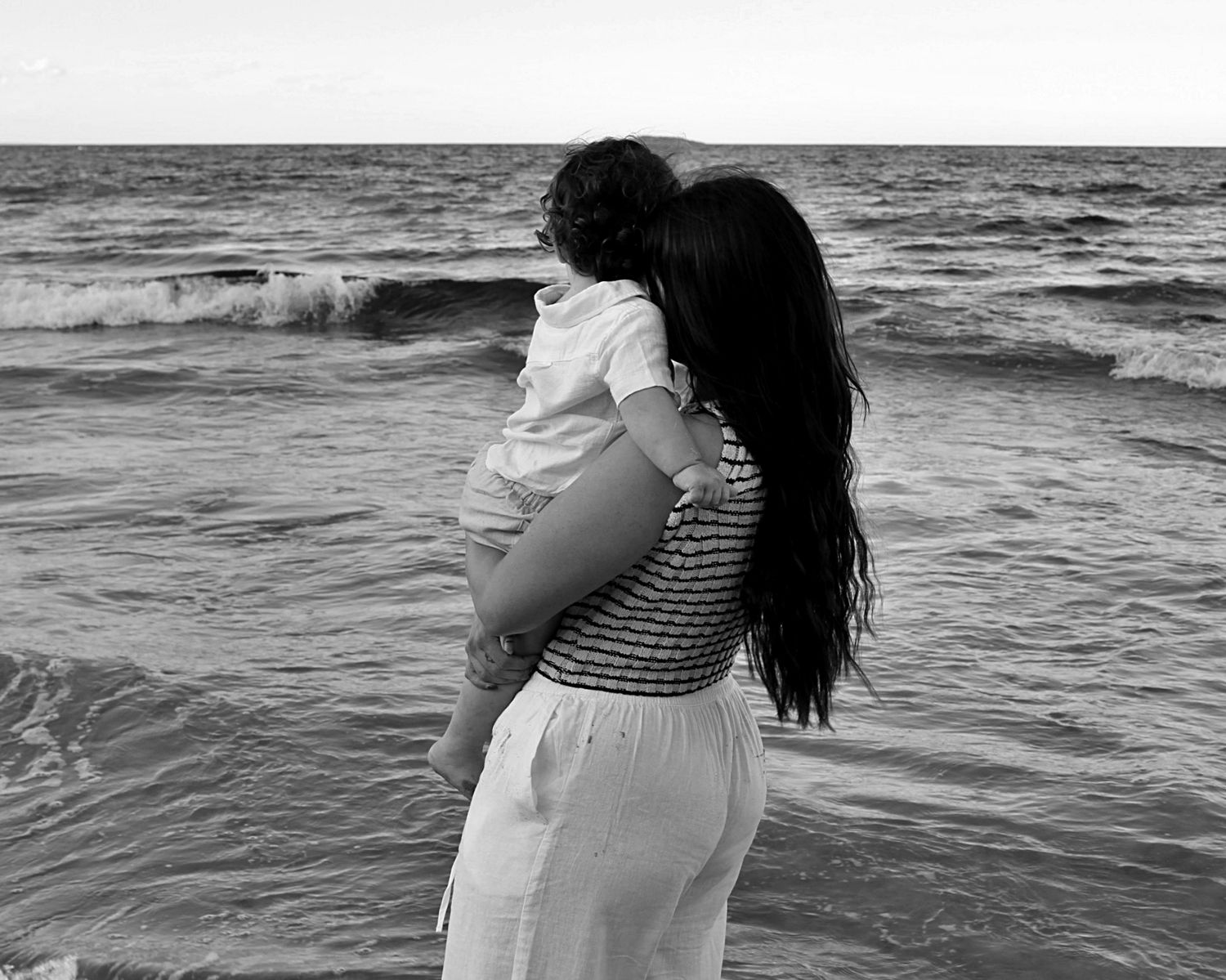 Mother holding her son on the shoreline during a beach motherhood session, looking out at the ocean, photographed from behind.