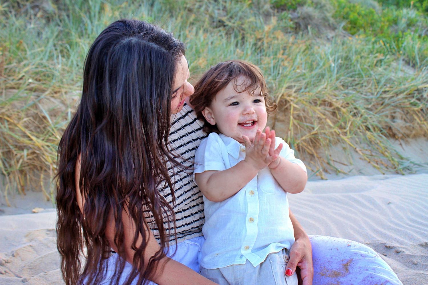 Beach motherhood session capturing a mother looking at her son while he claps excitedly on the sand.