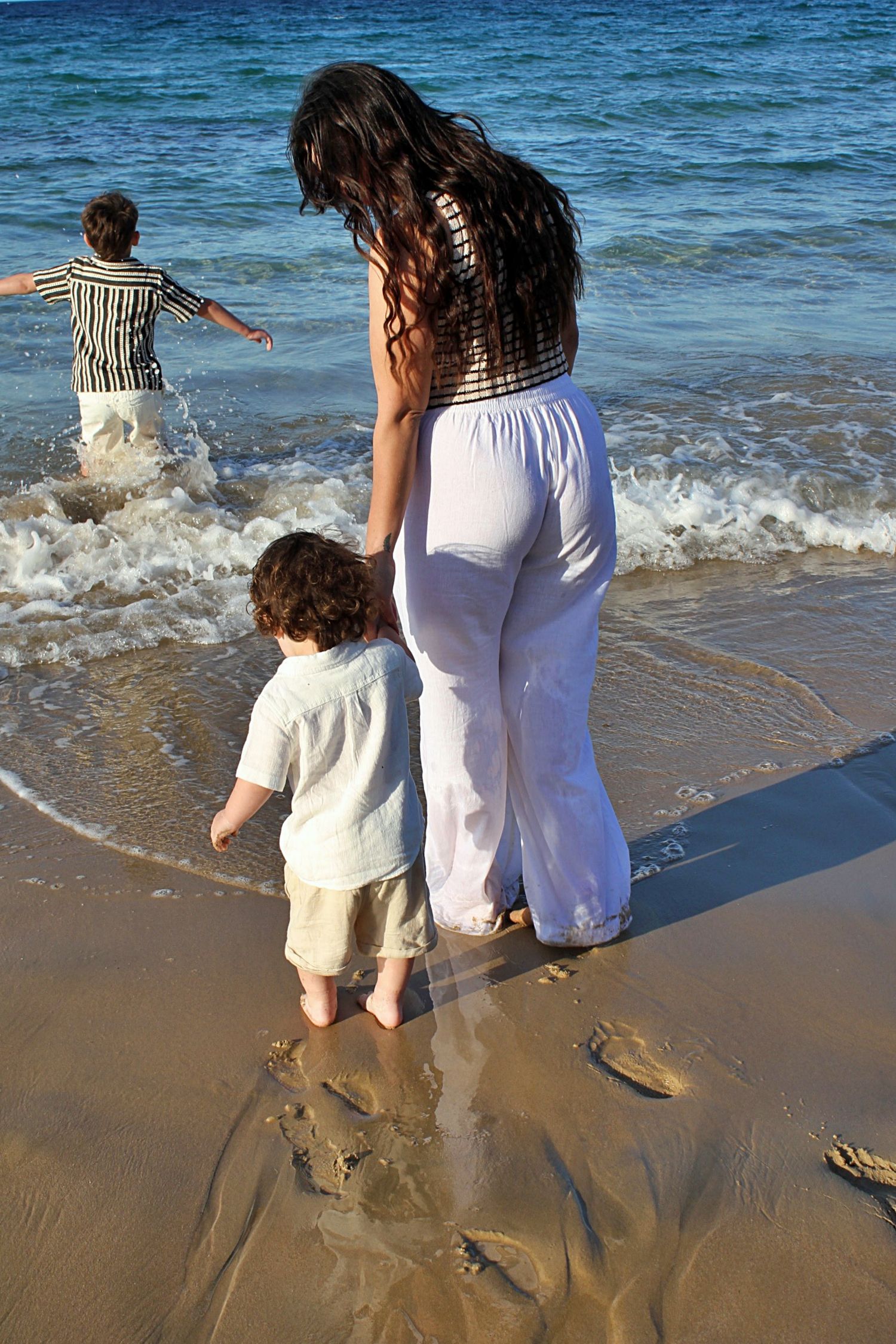Beach motherhood session showing a mother holding her son’s hand as they walk into the ocean together.
