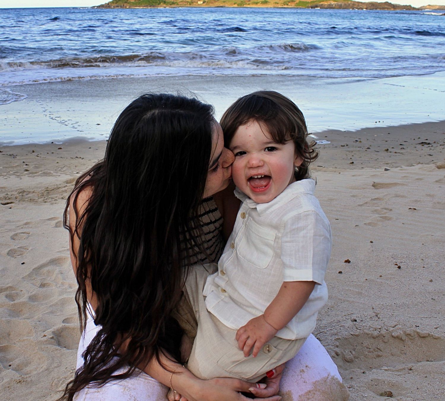 Candid motherhood session capturing a mother holding her son close while kissing his cheek by the ocean