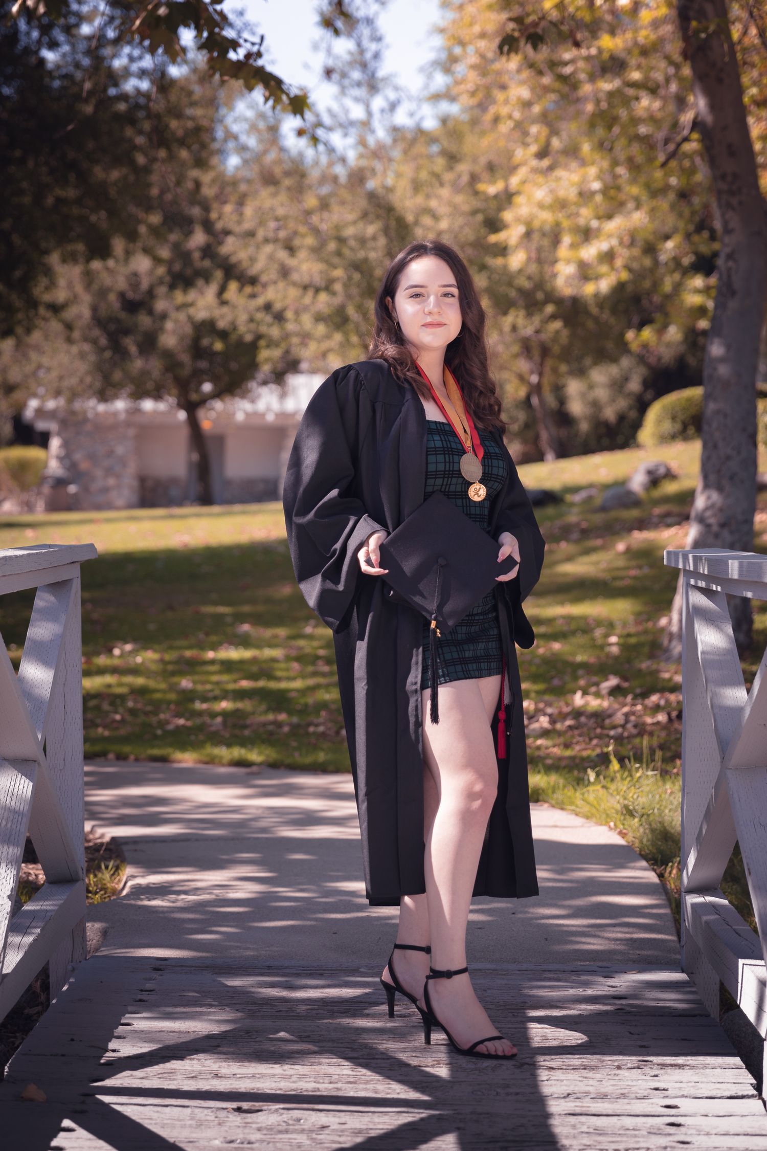 A person in a black coat poses on a walkway surrounded by autumn trees and fallen leaves on a sunny day.