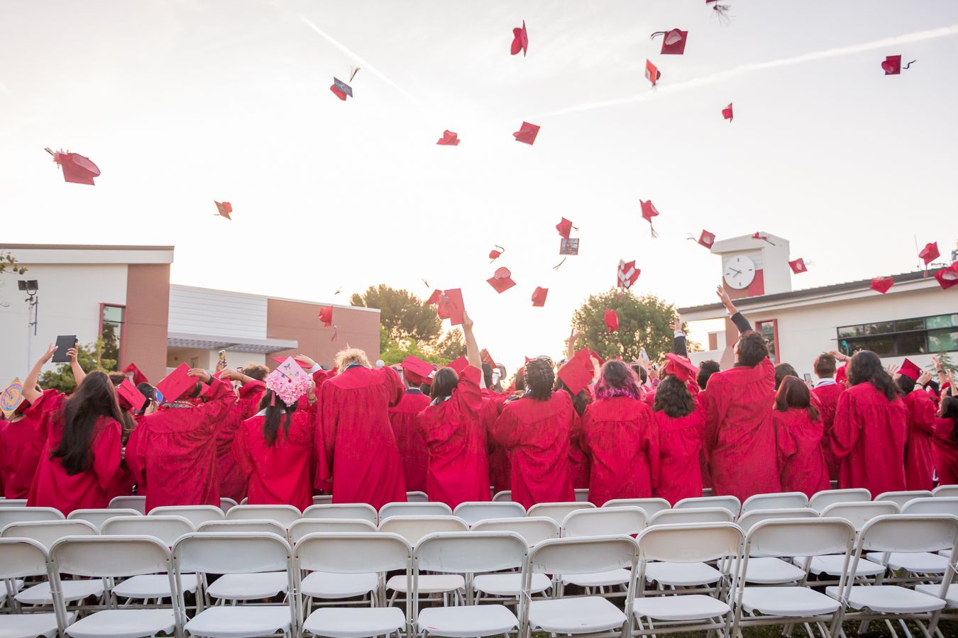 Graduates in red caps and gowns toss their mortarboards into the air during an outdoor graduation ceremony.
