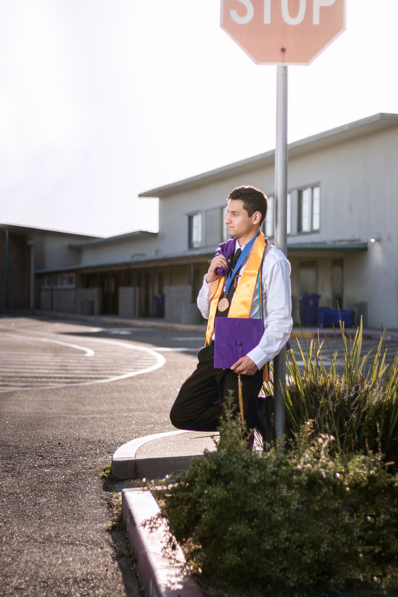 A person in purple and white sits on a curb near landscaping and a stop sign in a parking lot.