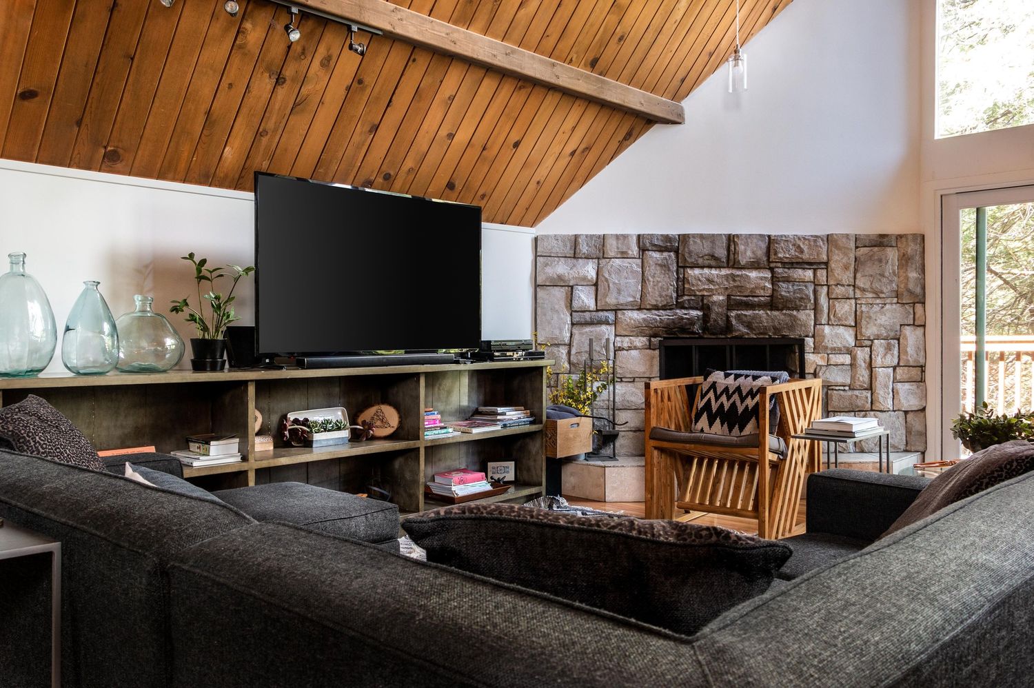 Modern living room with vaulted wood ceiling, stone fireplace, and large windows overlooking natural scenery.