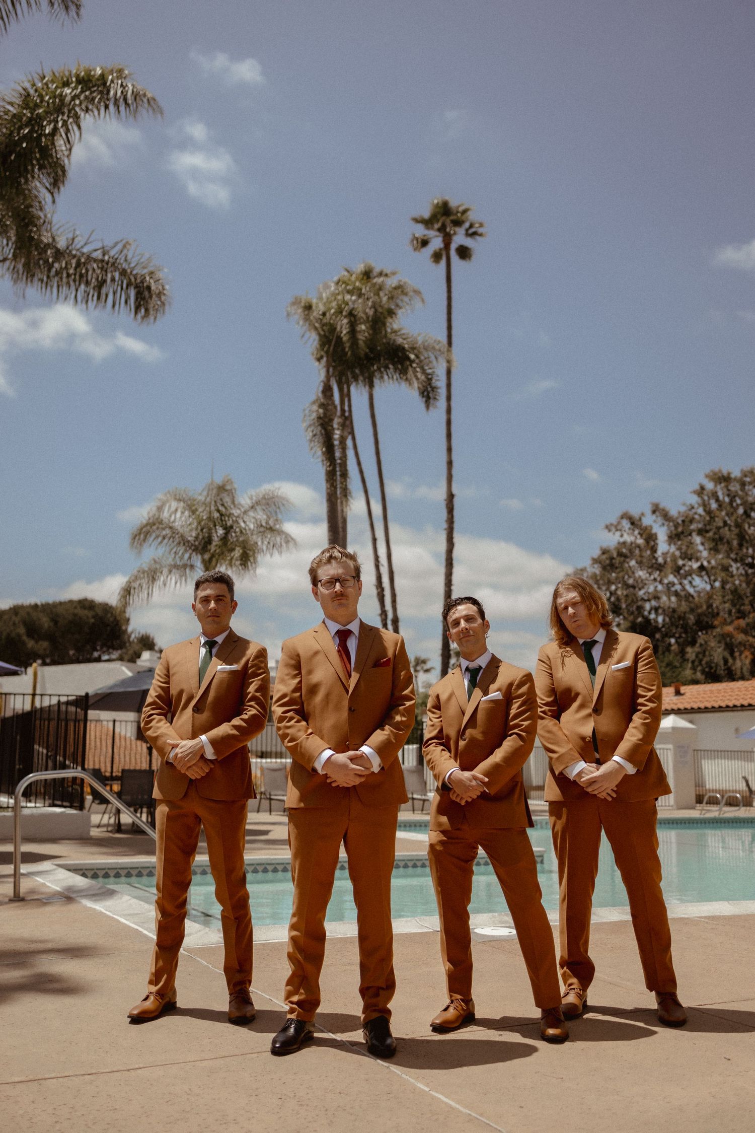 Groomsmen in matching tan suits pose beside a swimming pool with palm trees in the background on a sunny day.