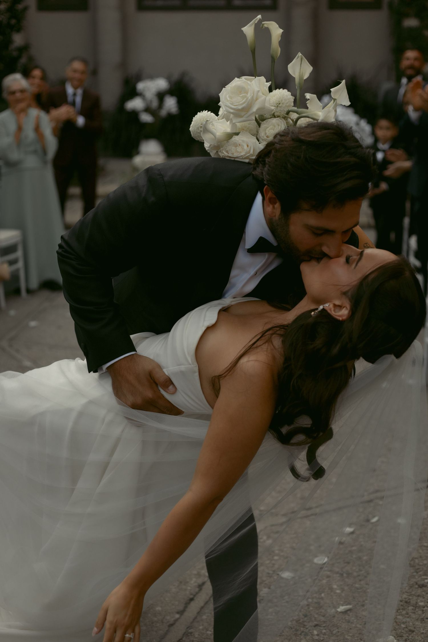 Groom dips bride for a romantic kiss at outdoor wedding ceremony with white floral bouquet and guests applauding.