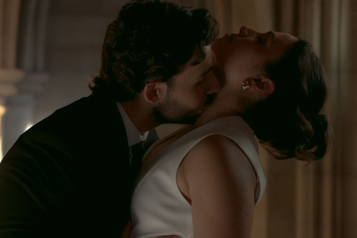 A couple shares a passionate kiss in a dimly lit, elegant setting, with the woman in a white dress.