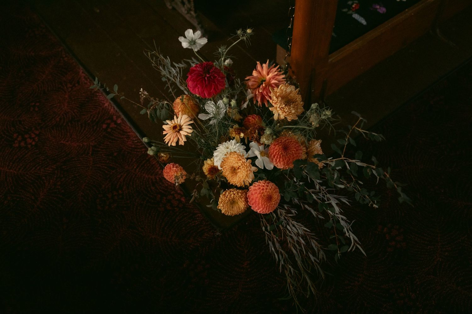 A moody floral arrangement featuring orange and peach blooms with wild greenery against a dark background.