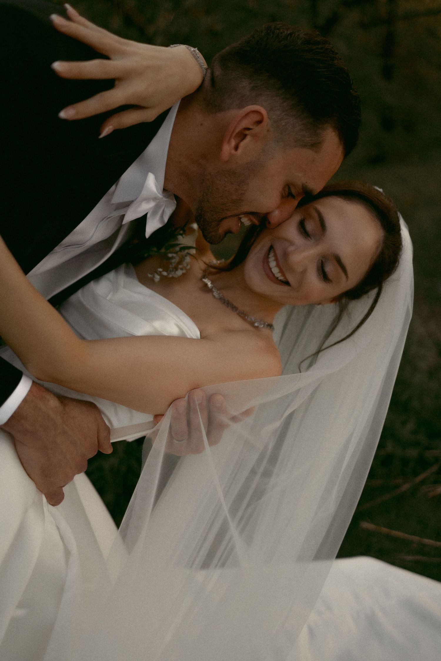 A groom in a black suit kisses his smiling bride wearing a white gown and veil on their wedding day.