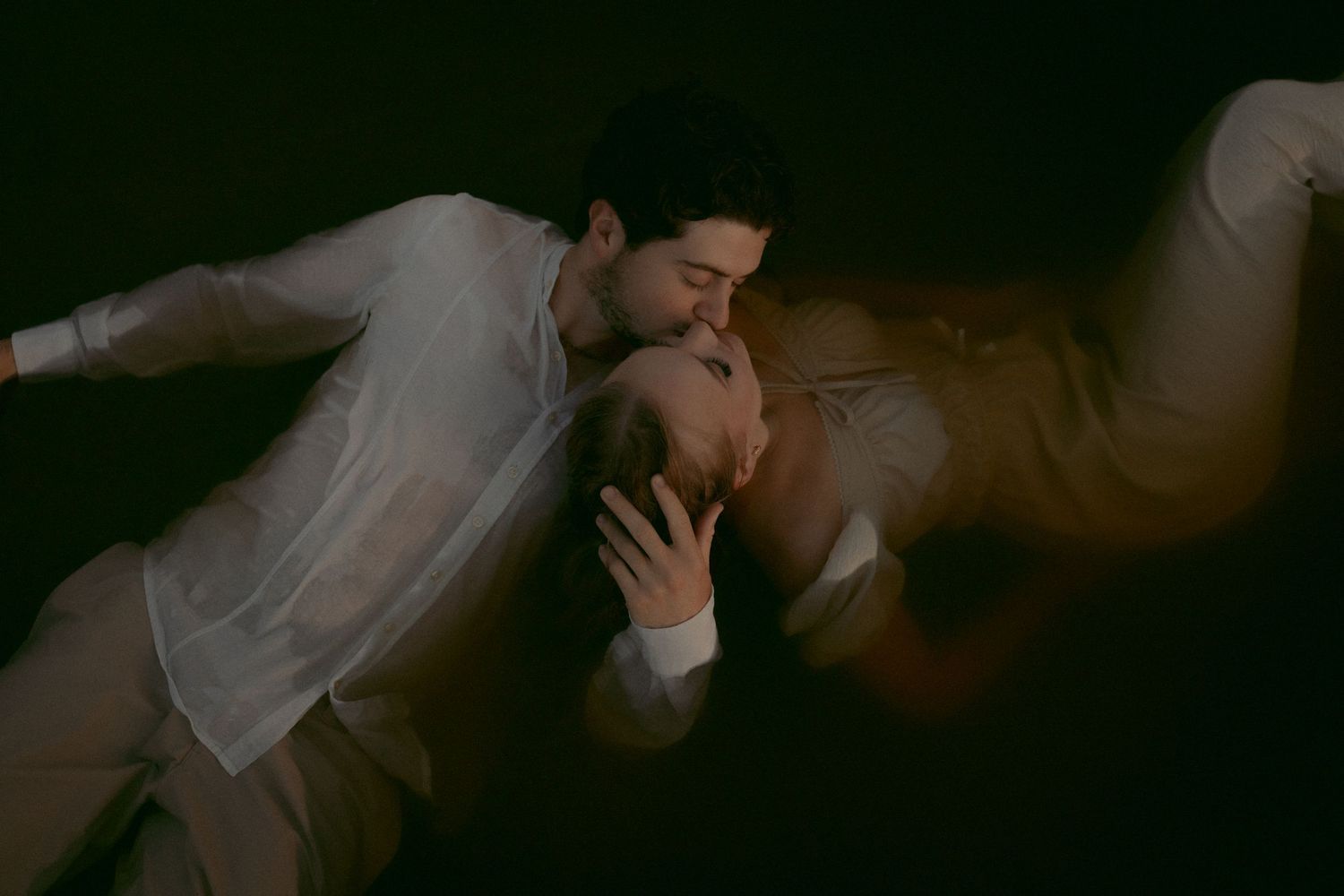 Romantic couple sharing a tender kiss in a dark, moody setting, wearing soft white and beige clothing.