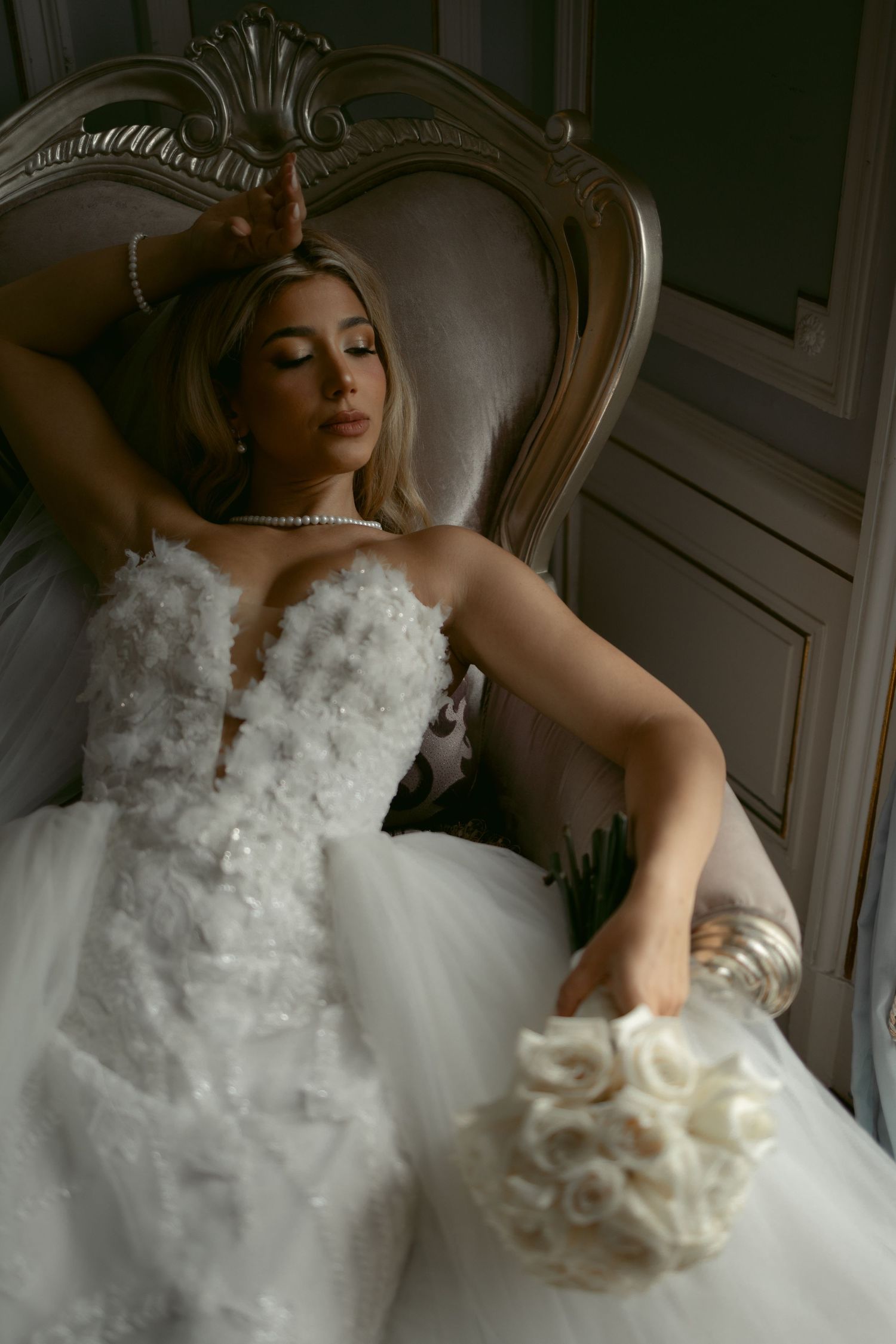 Elegant bride in strapless white tulle gown holding rose bouquet, reclining on ornate vintage chair.