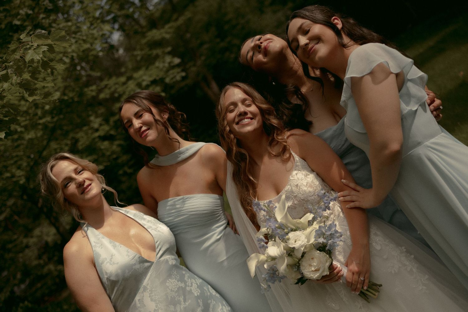 Bride holding blue and white bouquet smiles with four bridesmaids in light blue gowns outdoors.
