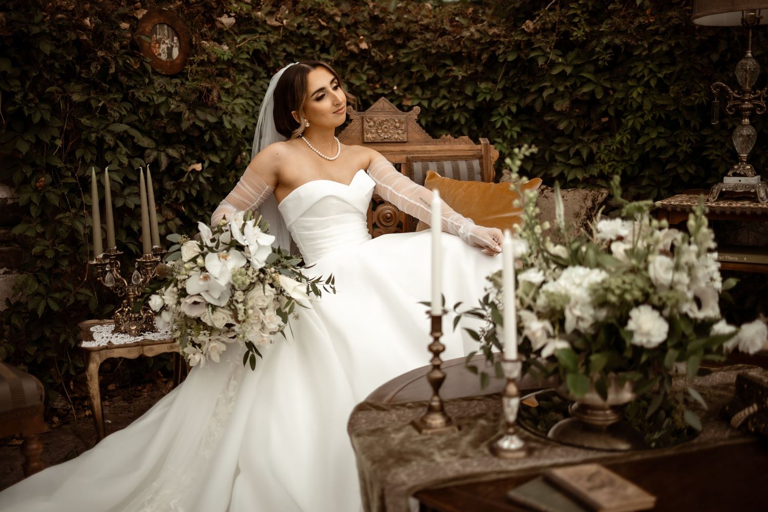A bride in a white strapless gown sits elegantly at a garden table decorated with floral arrangements and candles.