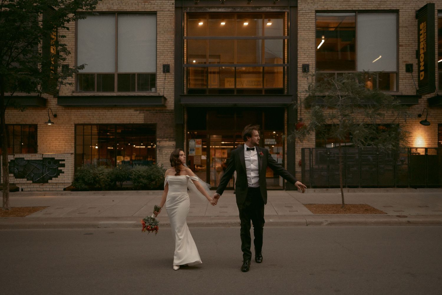 Bride and groom hold hands walking across urban street at dusk outside modern brick building with warm glowing windows.