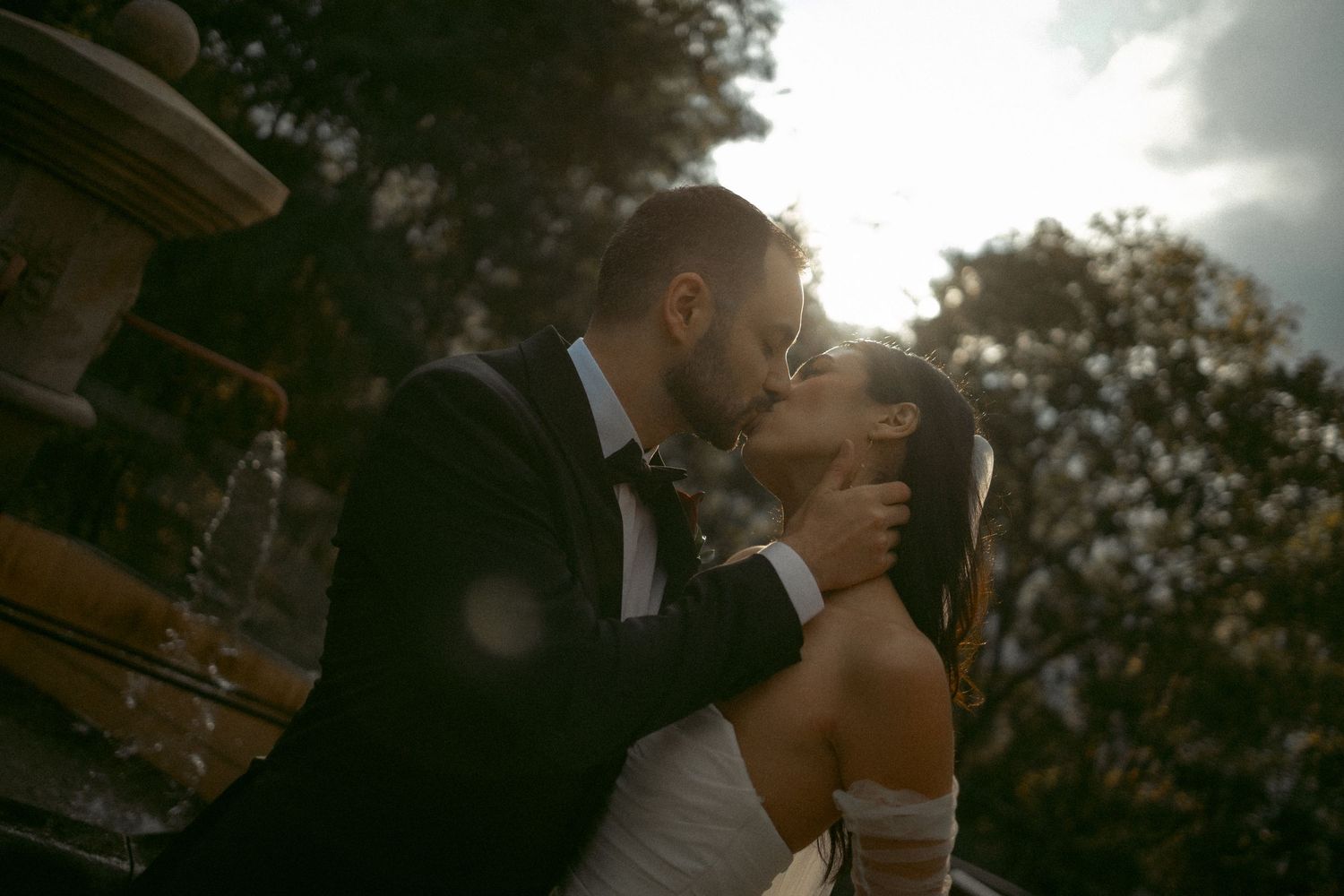 A couple shares a romantic kiss near a stone fountain, with golden sunlight filtering through trees on their wedding day.