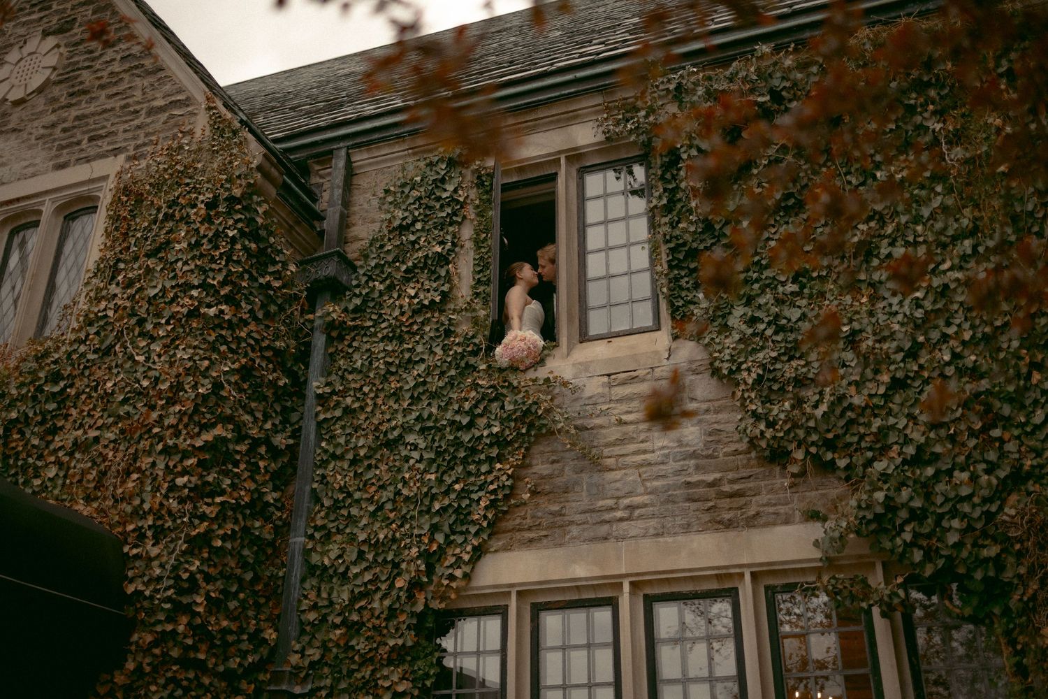 Couple posing in ivy-covered stone building window, romantic wedding photography at historic venue.