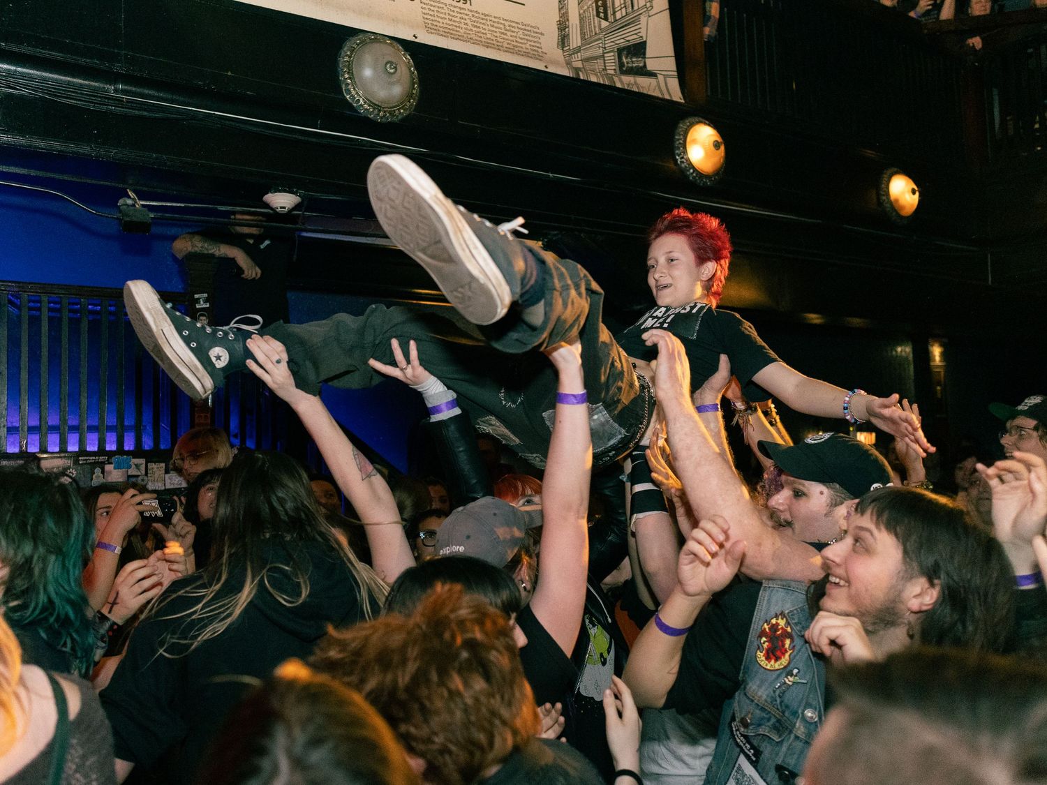 Full-color concert photograph of a person crowd surfing above an energetic audience during a live music performance.