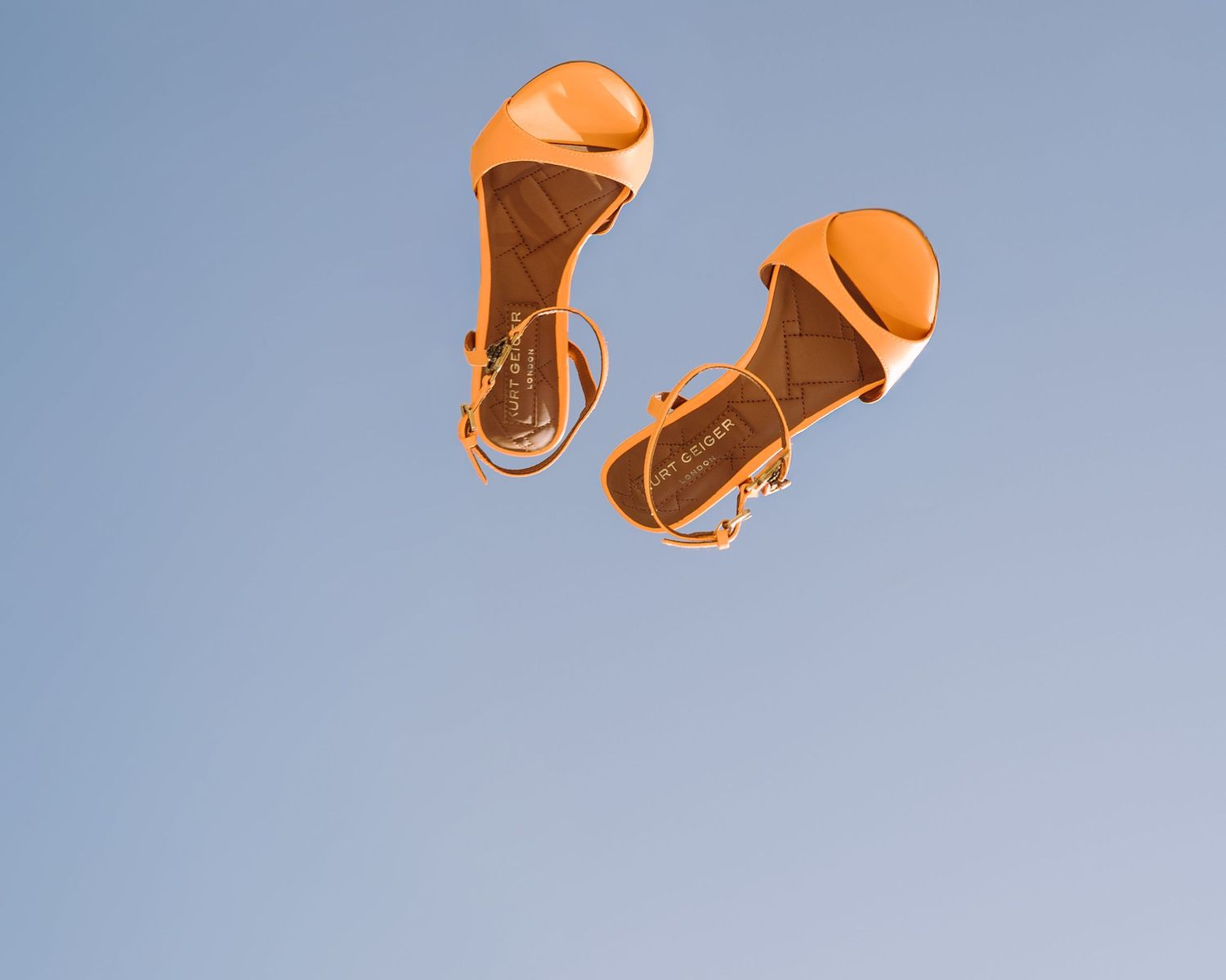 Orange bridal sandals photographed in mid-air against a clear blue sky, wedding detail