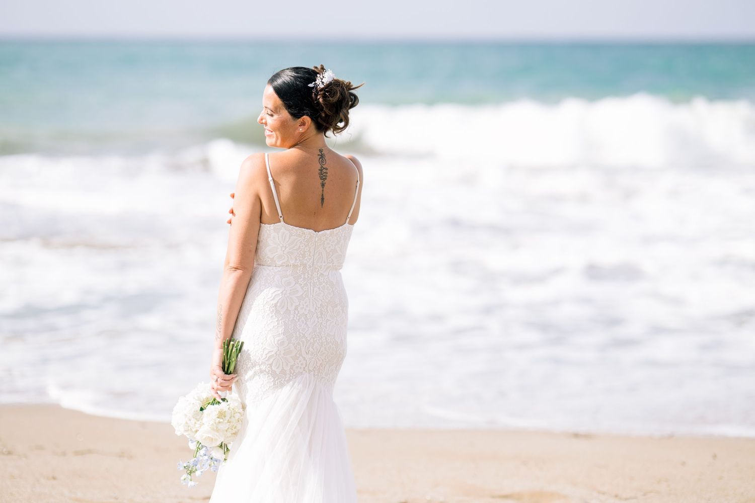 Bride with an elegant updo holding a white bouquet, looking out at the waves during a beach elopement in Crete