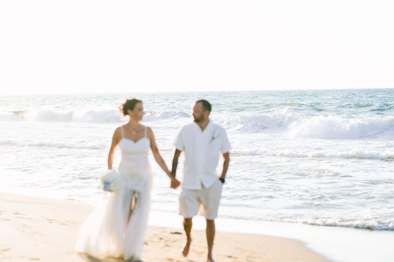 Couple walking hand in hand at the water's edge, the bride carrying a bouquet, beach wedding in Crete