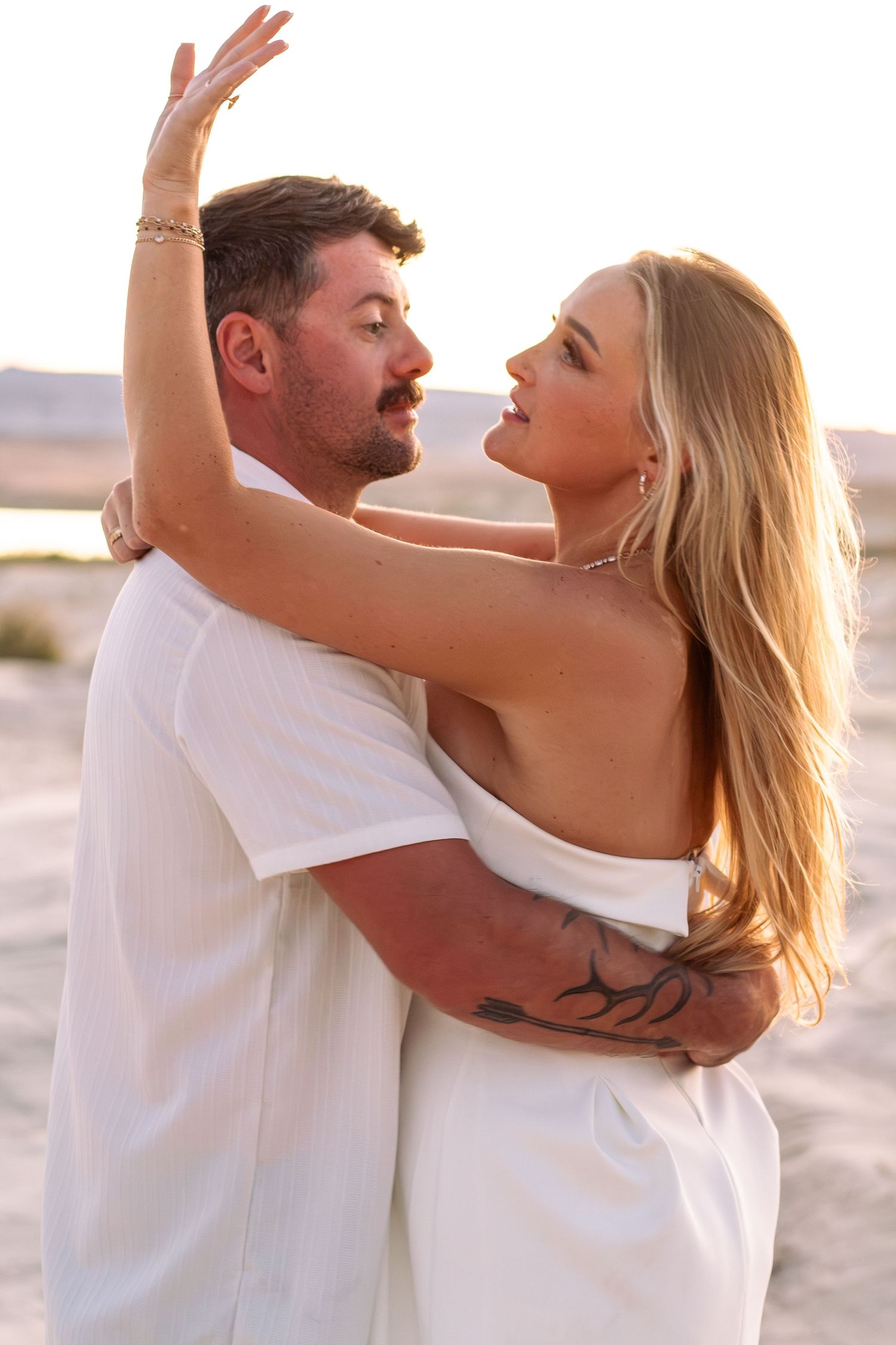 Couple in white outfits embracing on a beach at sunset, woman with blonde hair.