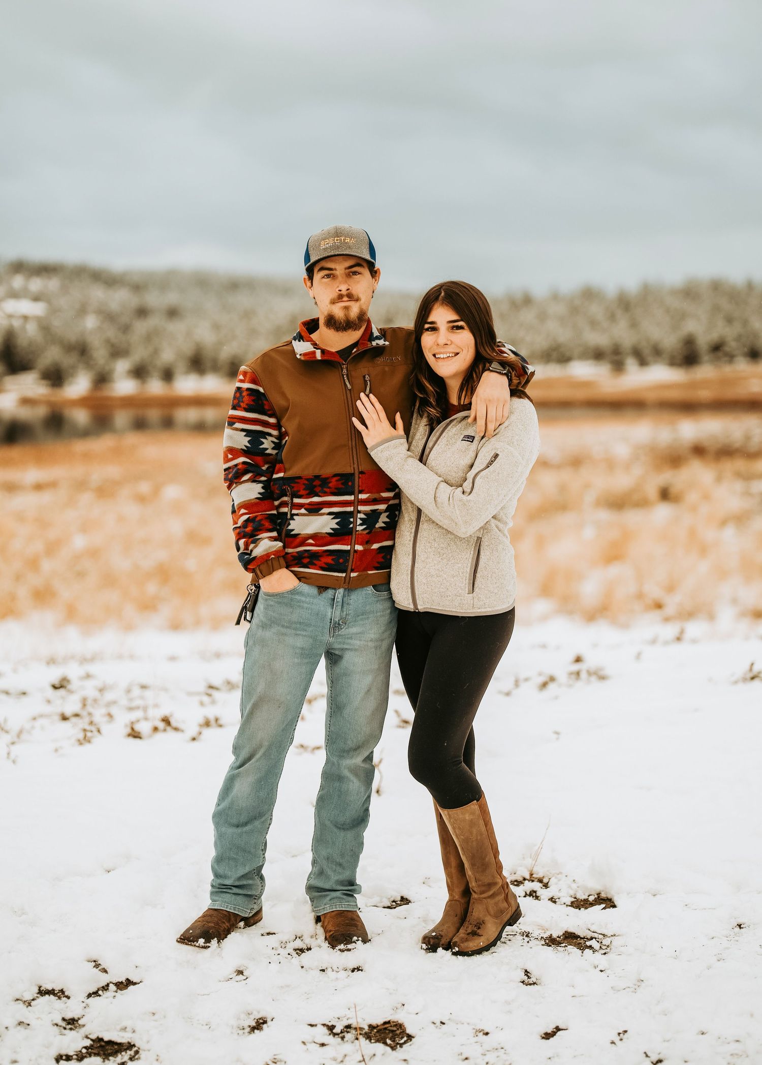 Couple posing together in snowy field, man in patterned sweater and cap, woman in beige jacket and brown boots.