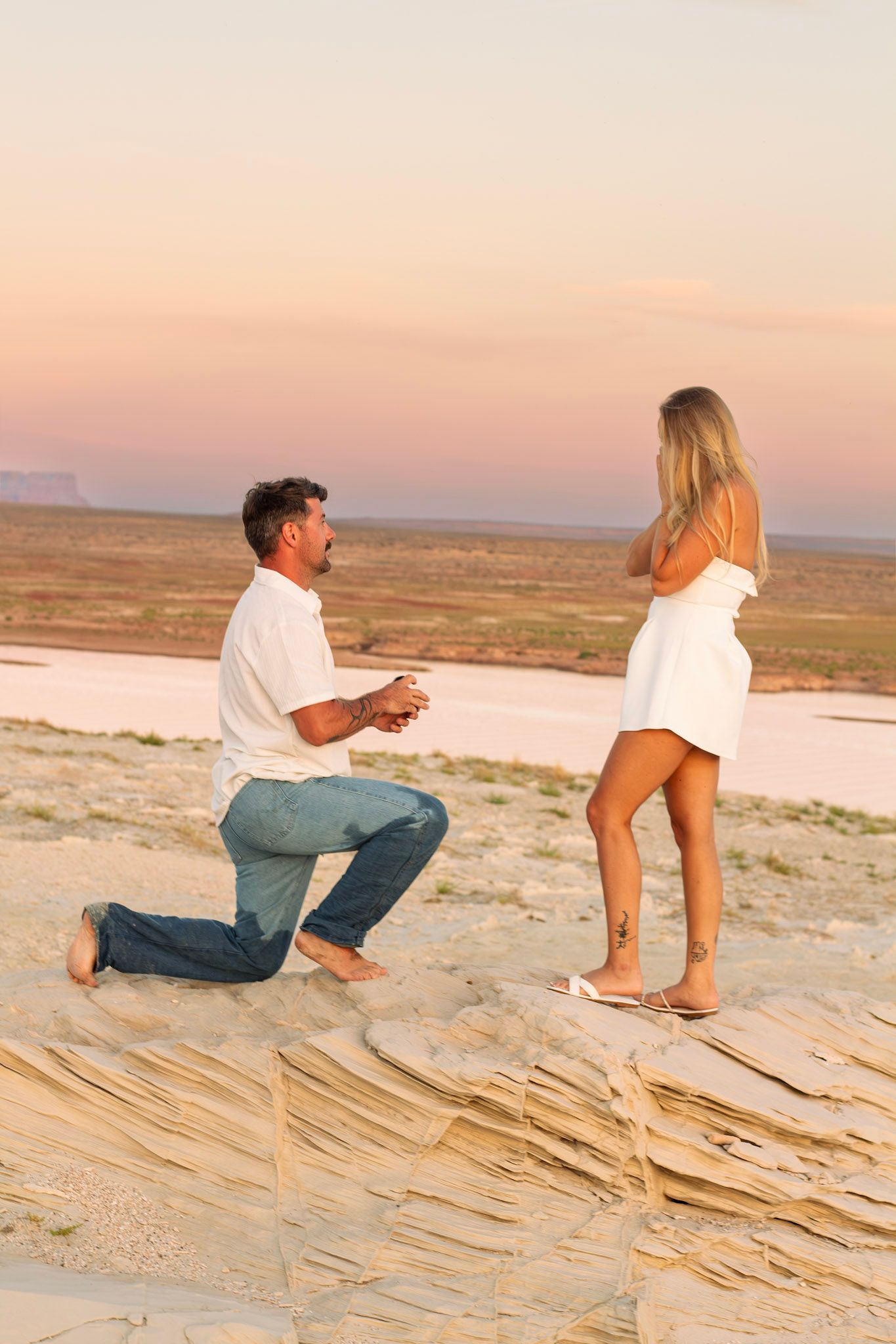A man proposes on one knee to a woman in a white dress on sandy cliffs at sunset.