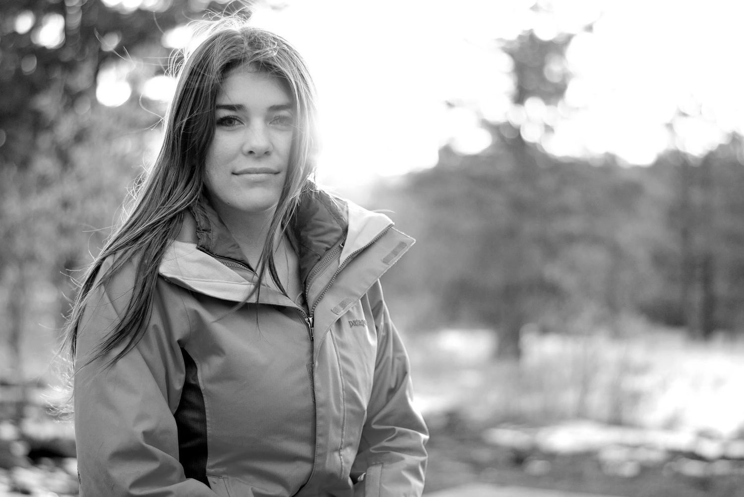 A young woman in an outdoor jacket poses in a black-and-white forest setting with a soft background.
