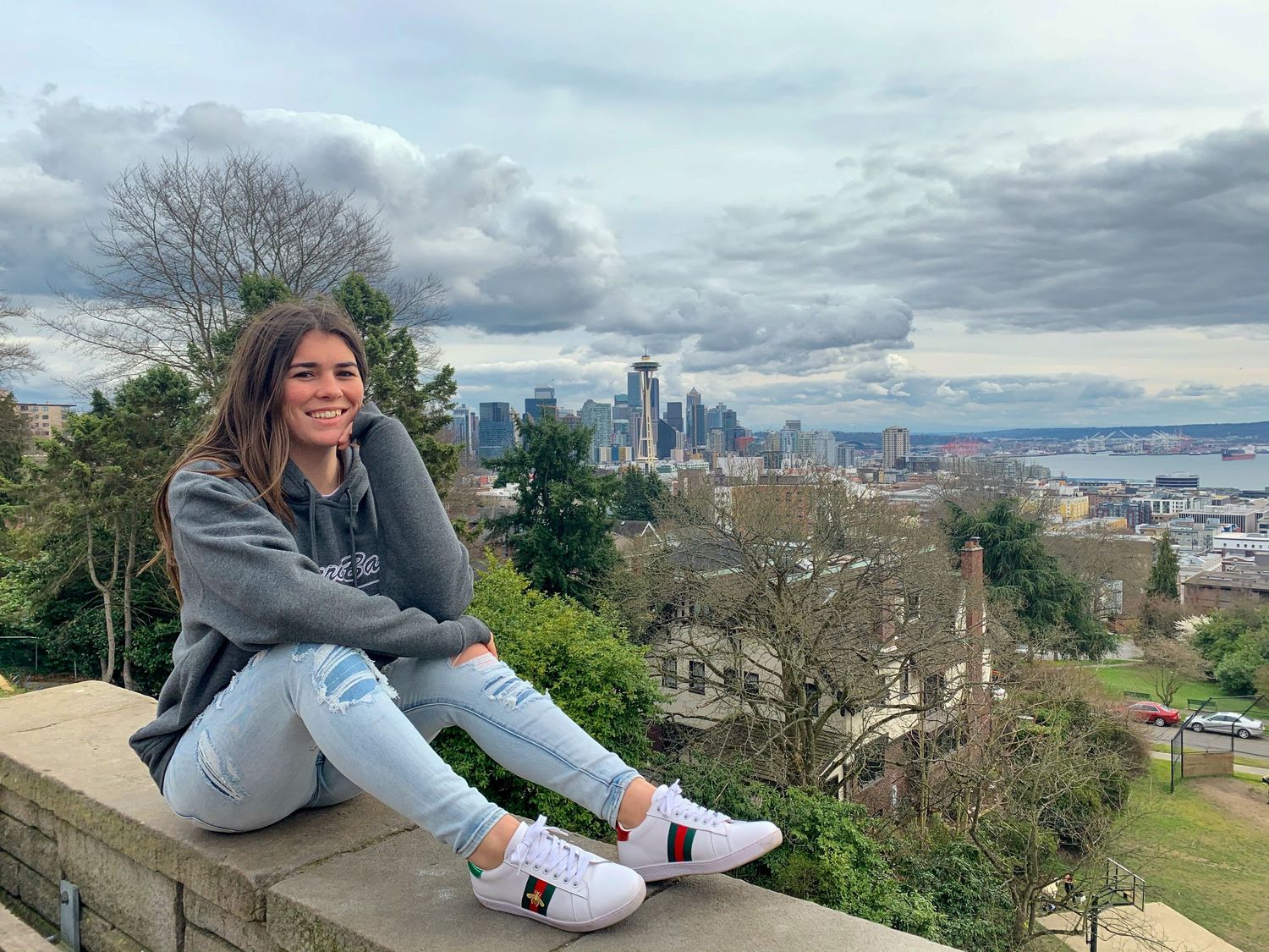 Young woman sitting on a stone wall with Seattle city skyline and Puget Sound in the background.