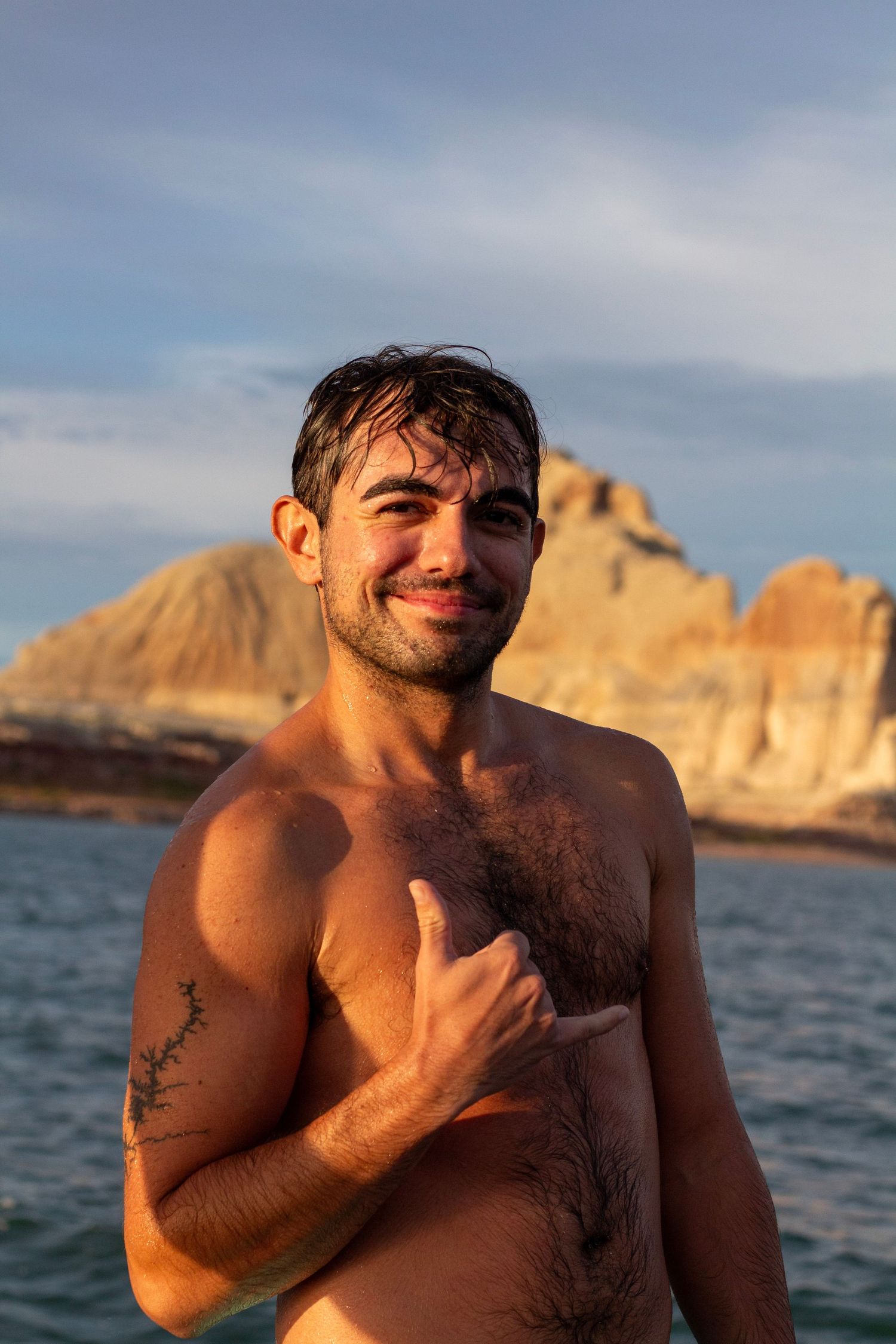 Shirtless man with wet hair smiling outdoors near rocky cliffs at golden hour.