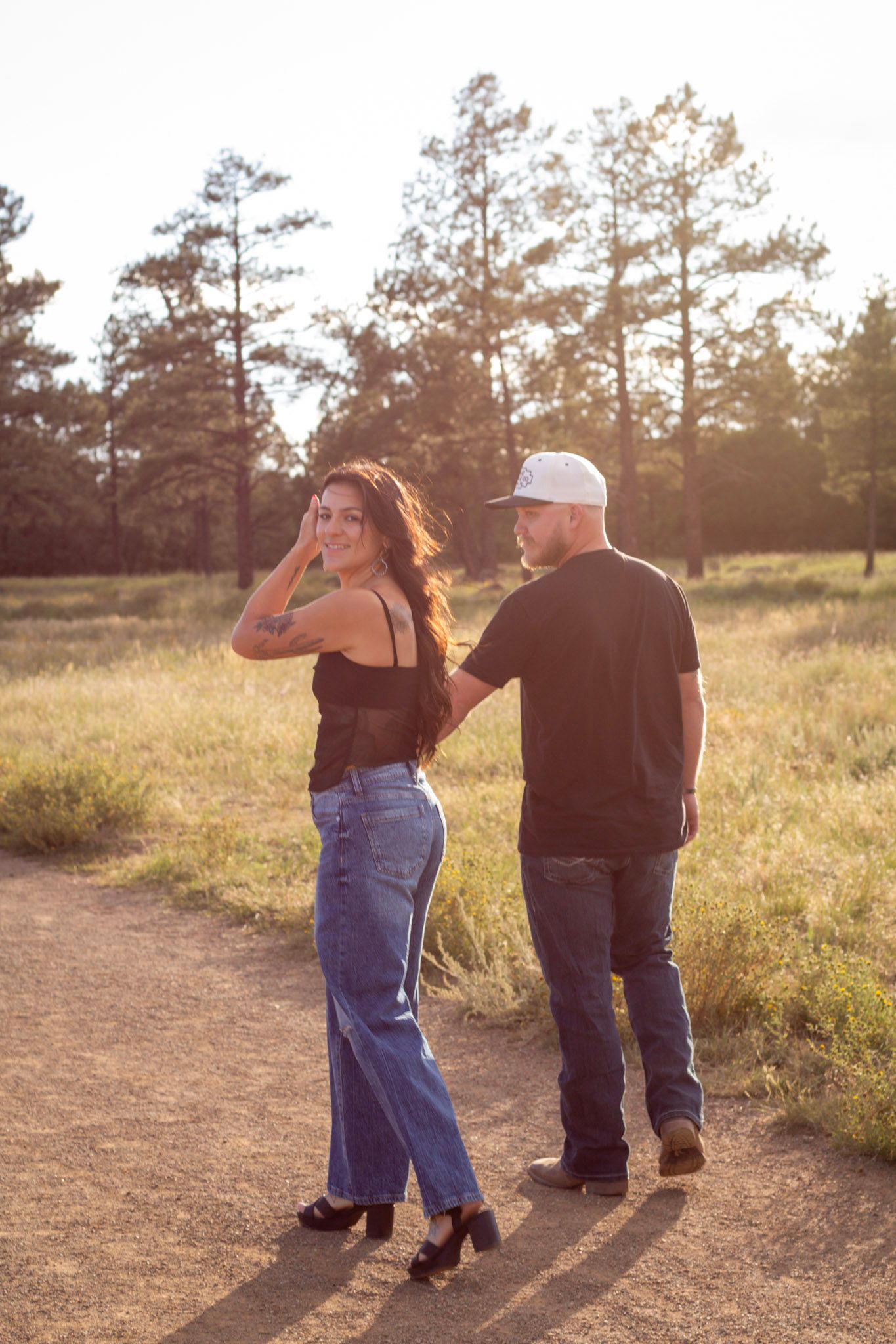 Couple walking on a dirt path at golden hour, woman in black top and jeans smiling, man in black tee and cap, pine trees behind.