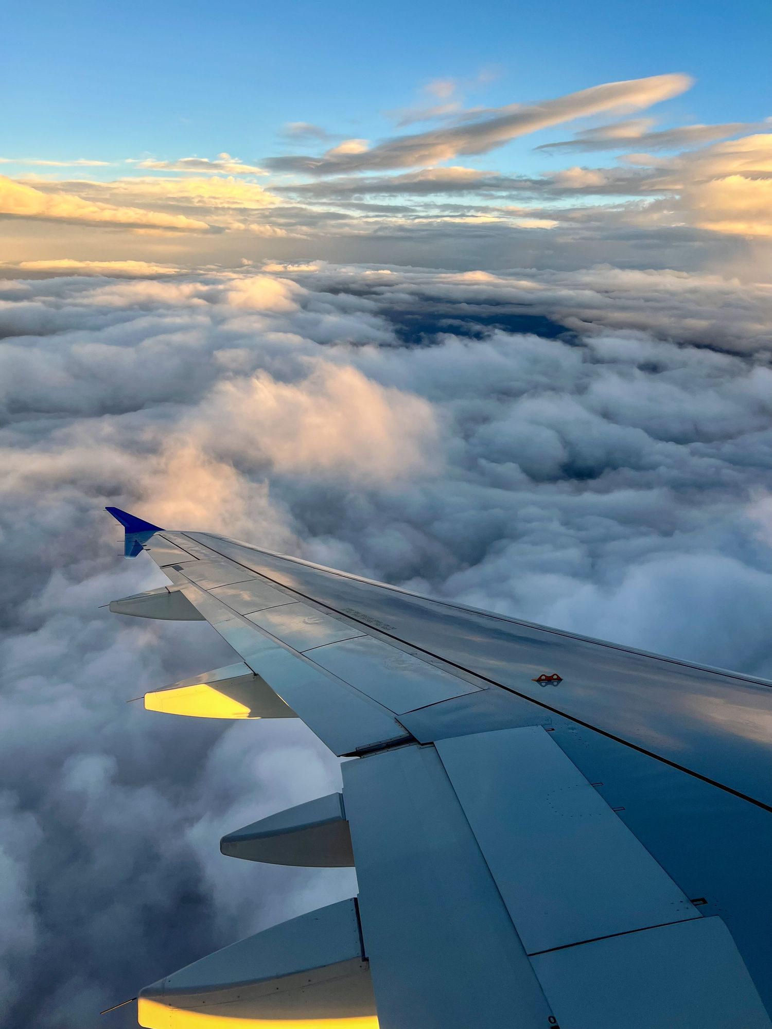 Airplane wing soaring above dramatic clouds at golden hour sunset, aerial view from passenger window seat.