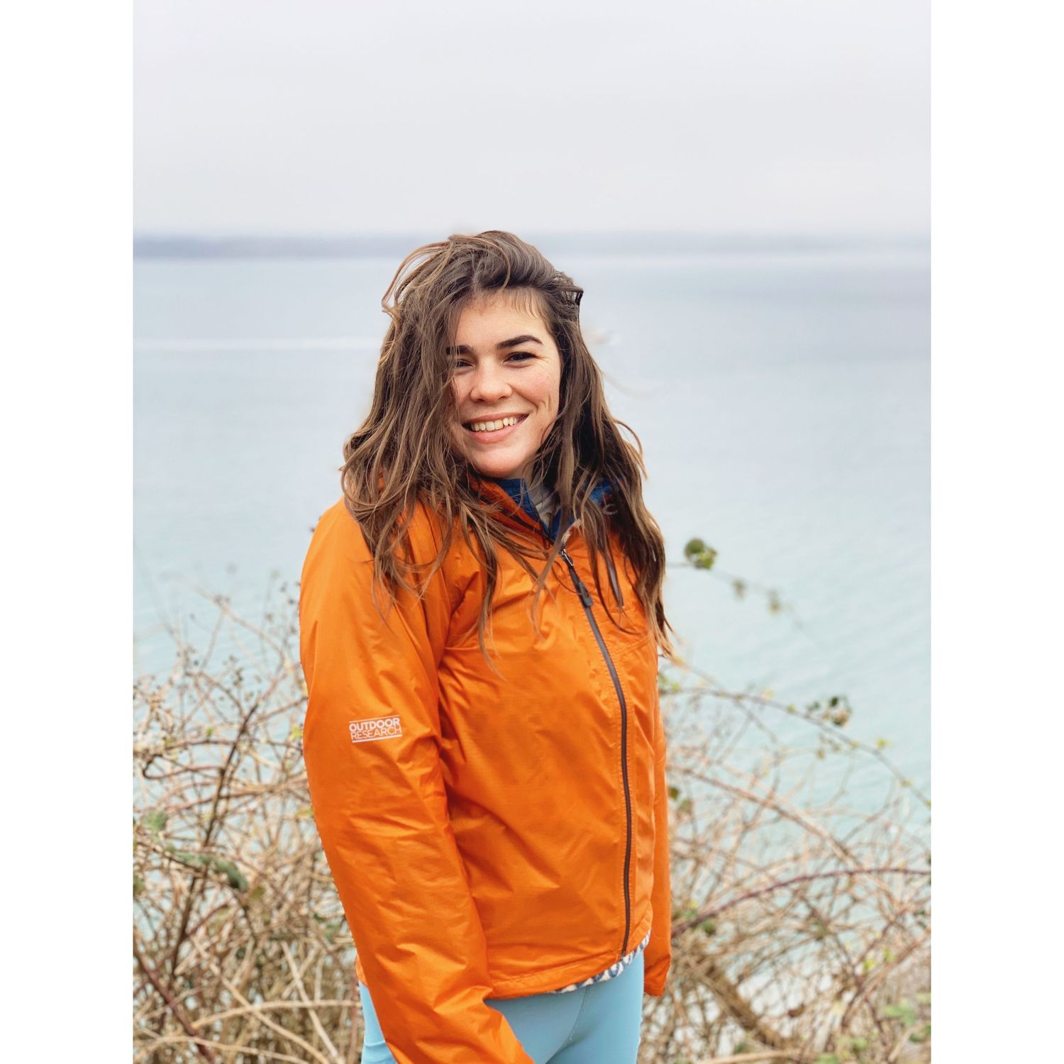 Smiling woman in an orange jacket standing outdoors with the ocean and cloudy sky in the background.