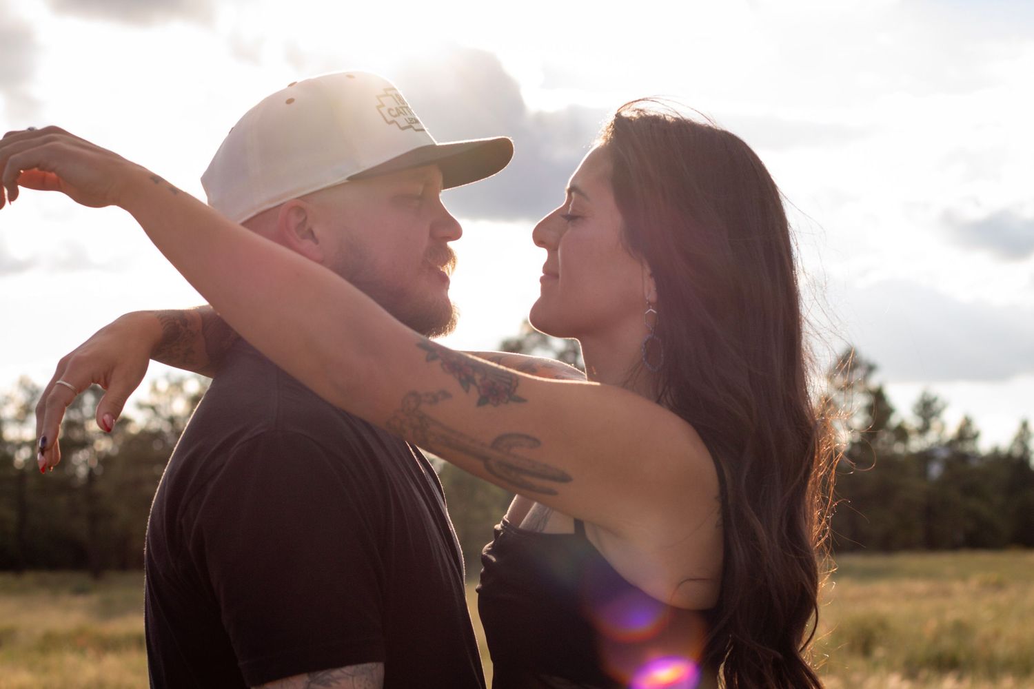 A couple shares a romantic moment outdoors, the woman wrapping her arms around a bearded man in a cap, backlit by sunlight.