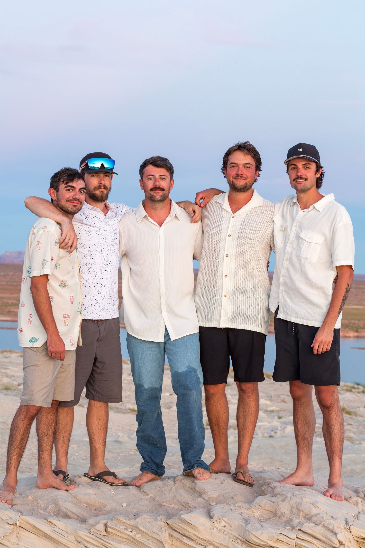 Five young men posing together on a sandy beach at sunset, wearing casual summer clothing and smiling.