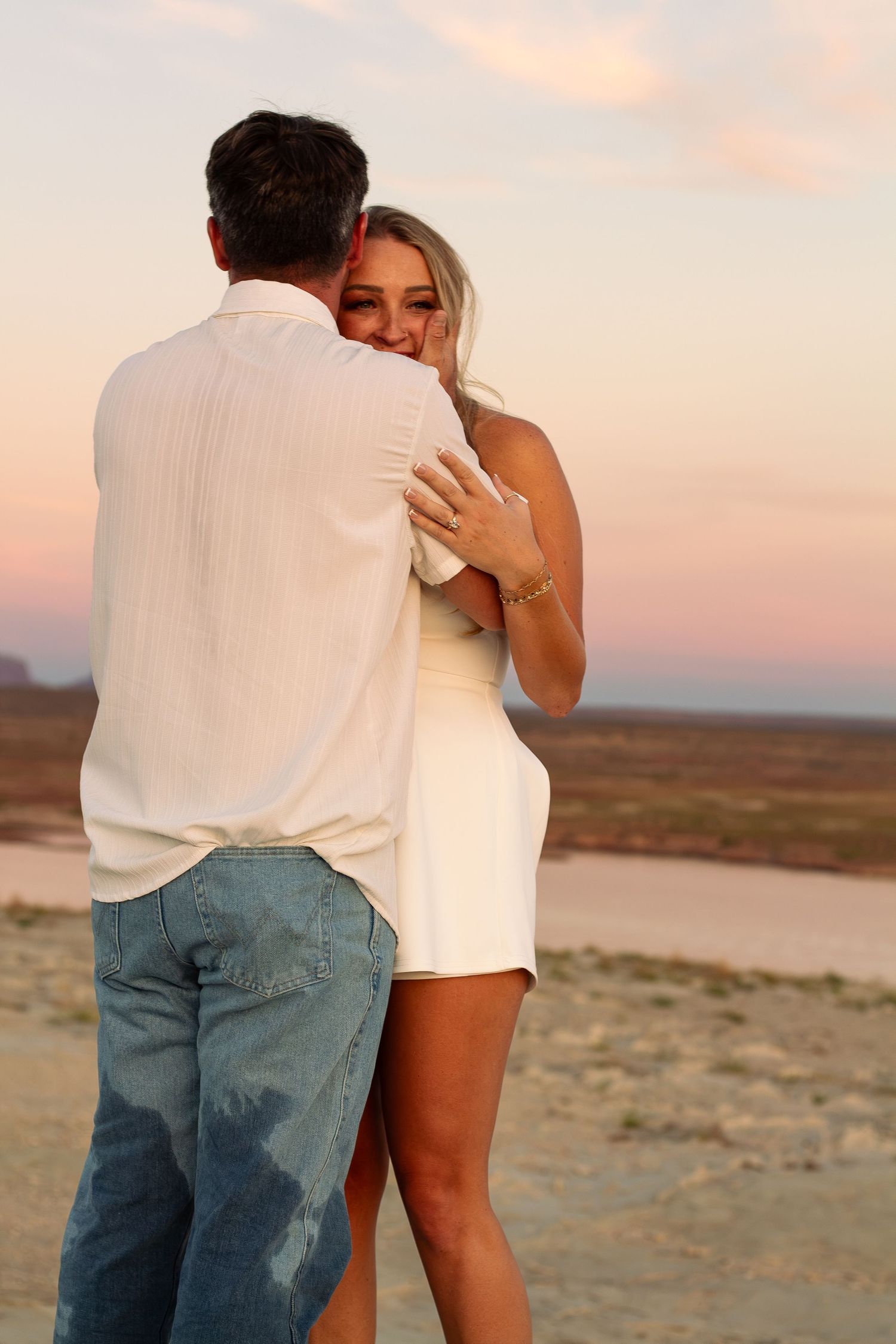 A couple embracing romantically on a sandy beach at sunset, both wearing white outfits, with a colorful pink sky backdrop.