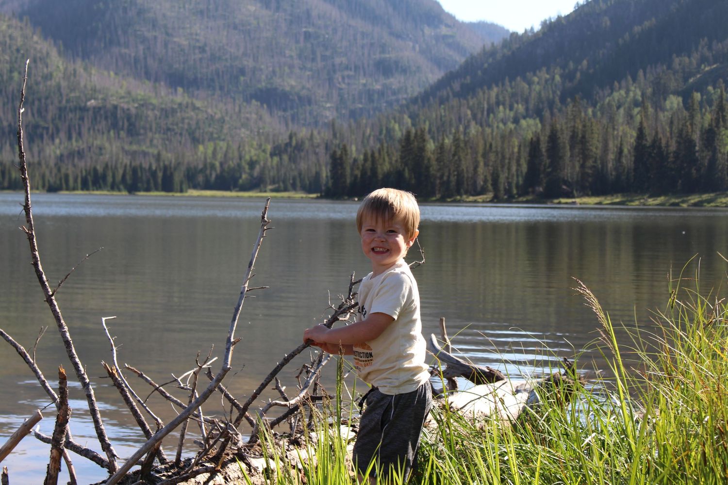A young boy is smiling by a mountain lake shore with forested hills in the background on a sunny day.