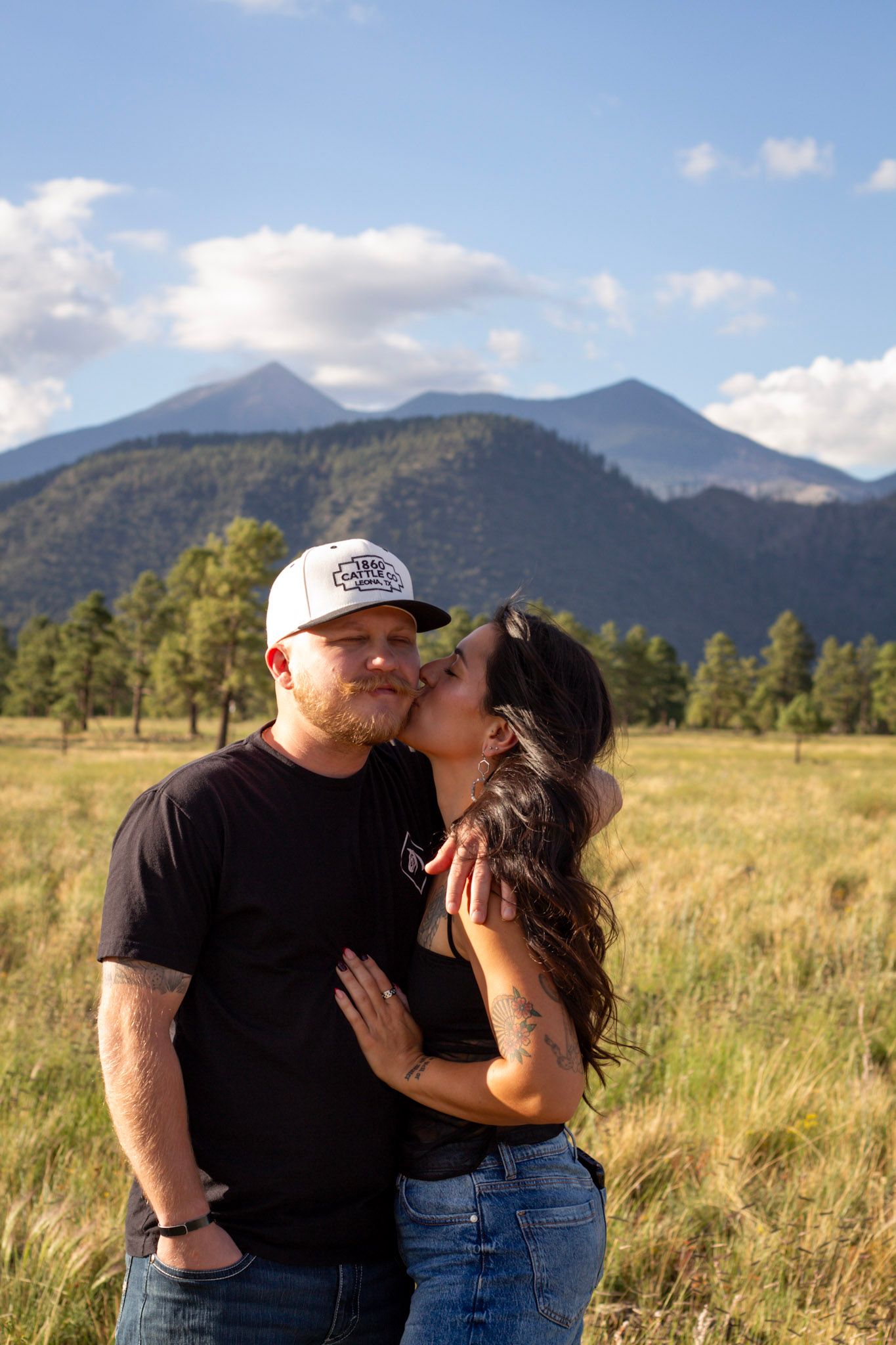 A couple shares a romantic kiss in a mountain meadow, with pine trees and peaks under a blue sky.