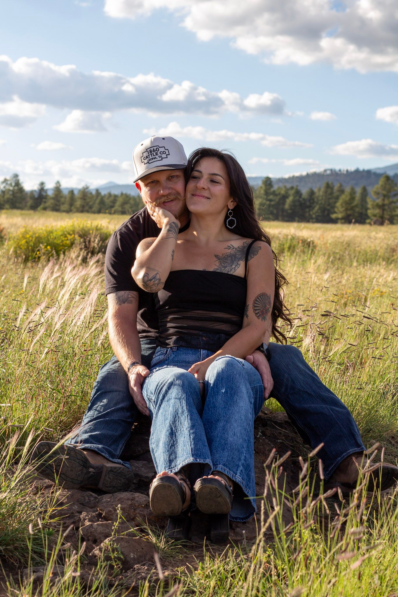 A couple sits together in a sunny meadow, the woman leaning back against her partner, both smiling contentedly.