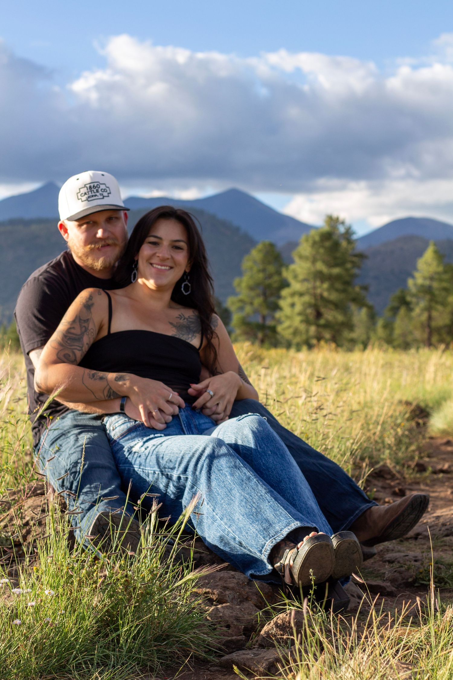 A couple sits together in a mountain meadow, the woman smiling in a black top and jeans, the man in a cap embracing her.