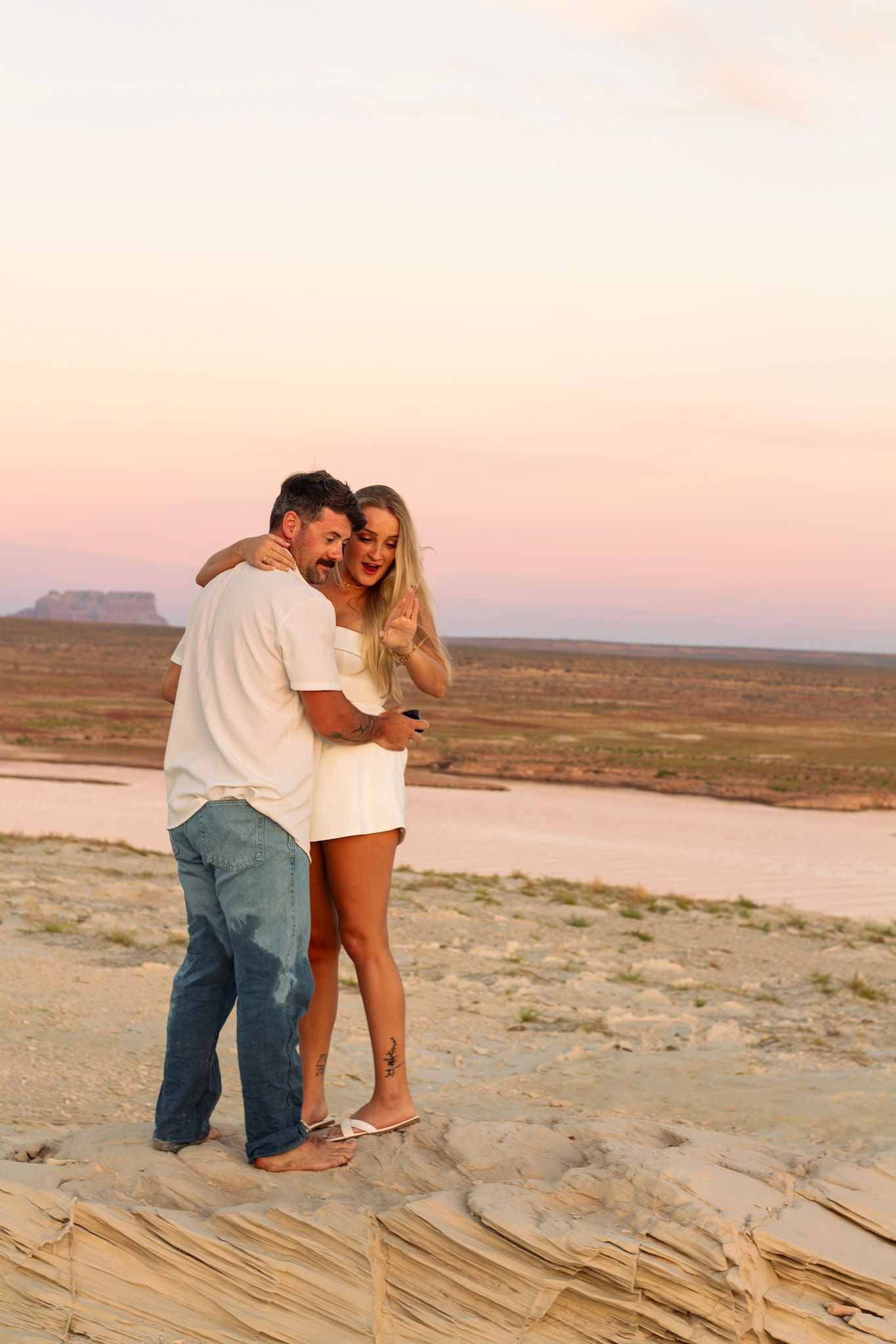 A couple embracing and smiling at sunset on a rocky desert landscape with a pink sky and a mesa in the background.