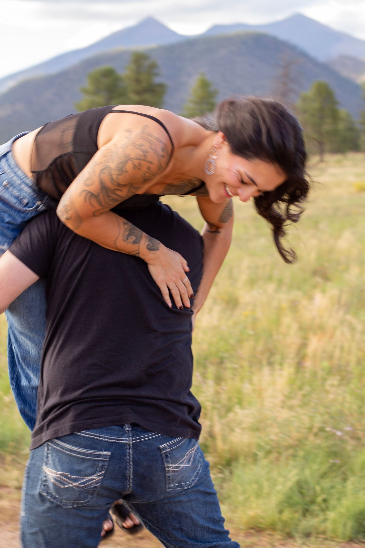 A woman laughing while being thrown over a man's shoulders, outdoors with mountain scenery in the background.