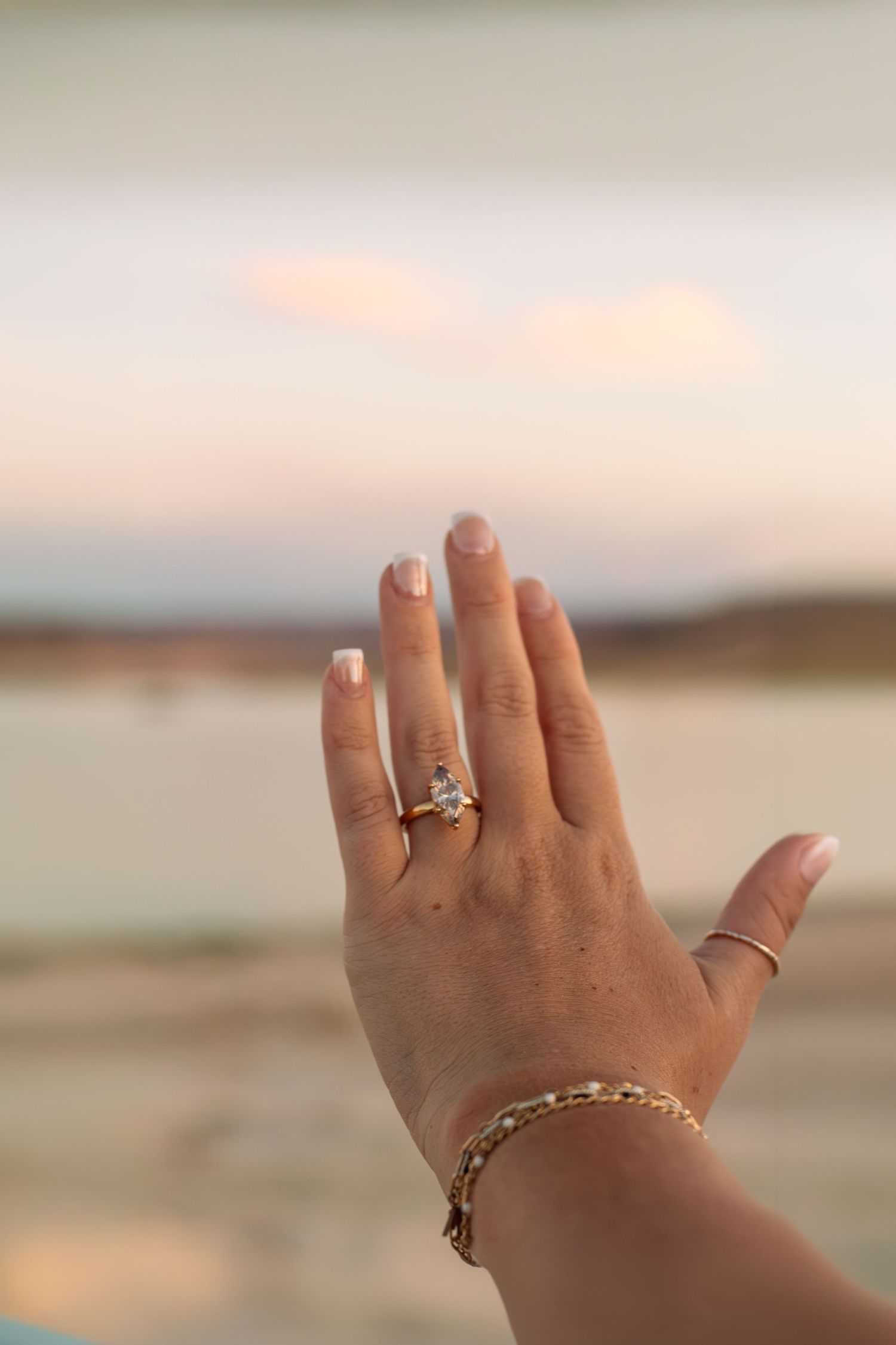Hand with diamond engagement ring, gold bracelet, and French manicure raised against a soft sunset beach backdrop.